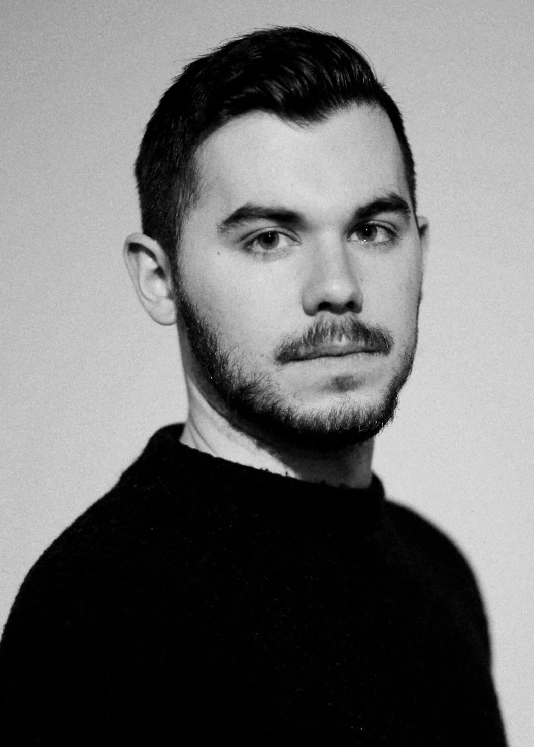 Black and white portrait of a young man with short dark hair, light facial hair, wearing a dark sweater, looking directly at the camera.