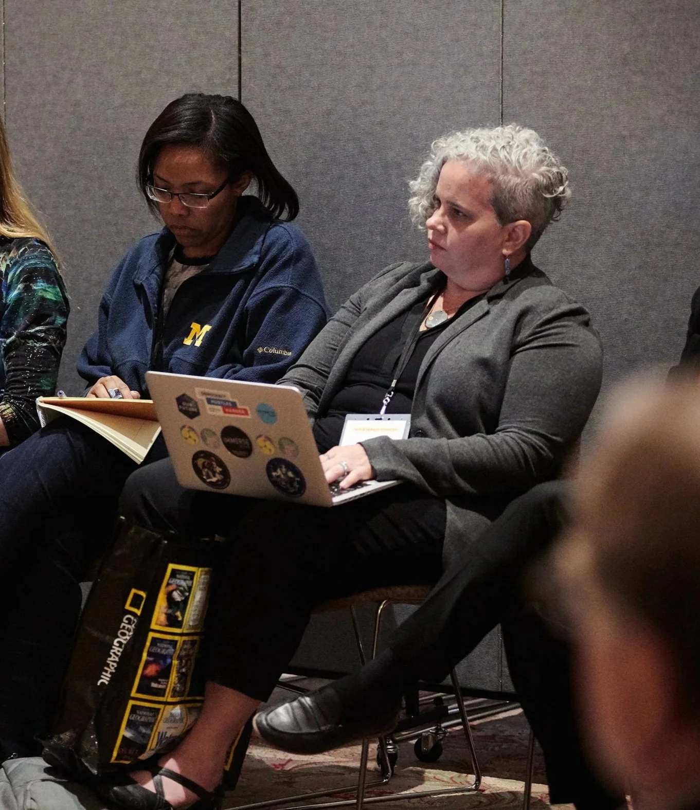 Group of people sitting at a conference or meeting, taking notes and using laptops, with gray walls in the background.