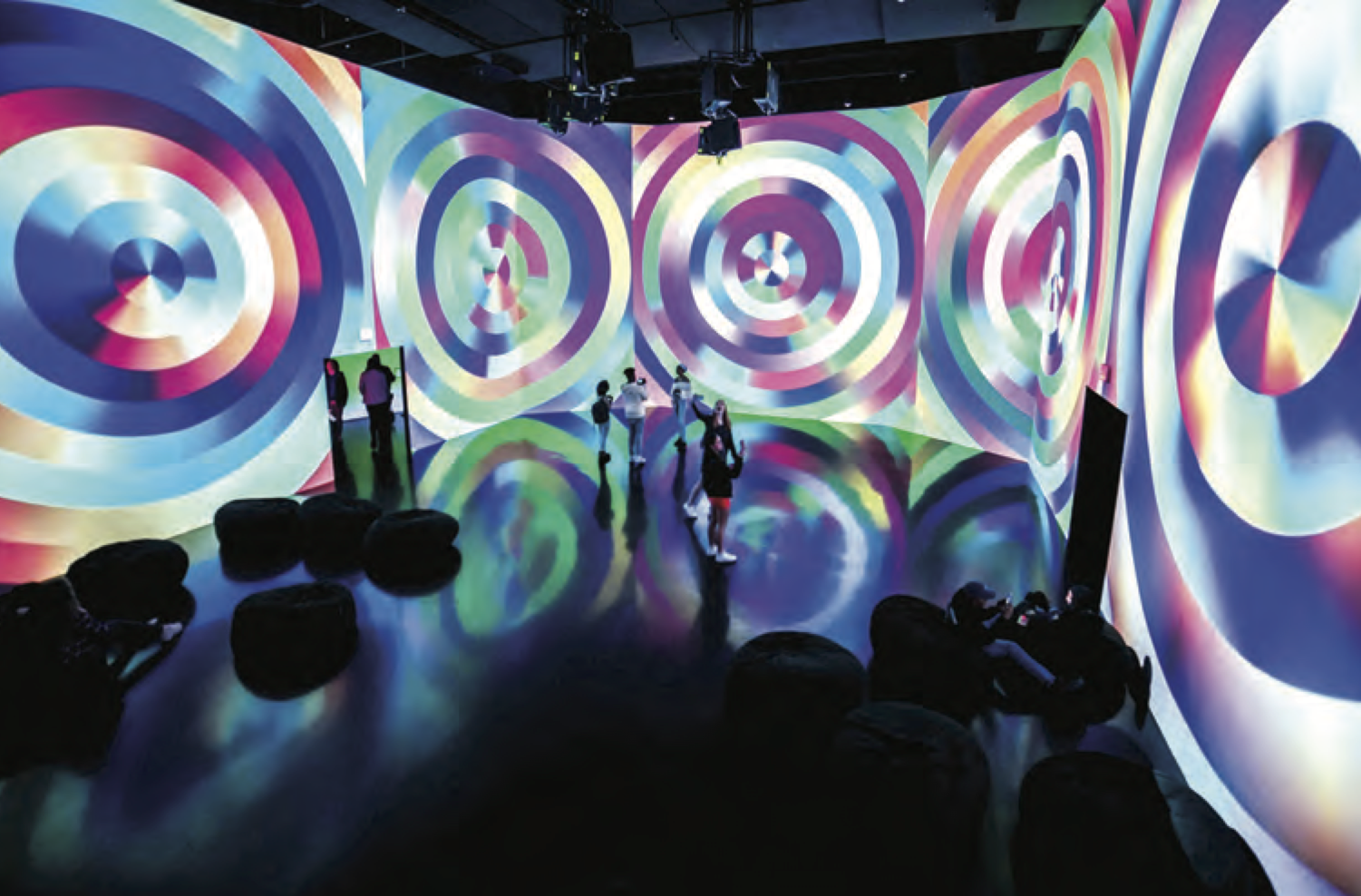 People observing a colorful light projection on walls and floor at a digital art exhibit.
