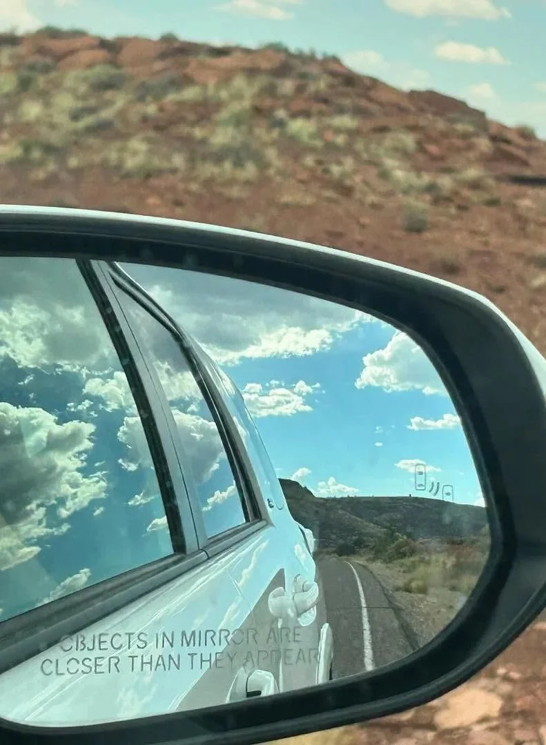 Side view mirror reflecting a cloudy sky and a road in a desert landscape.