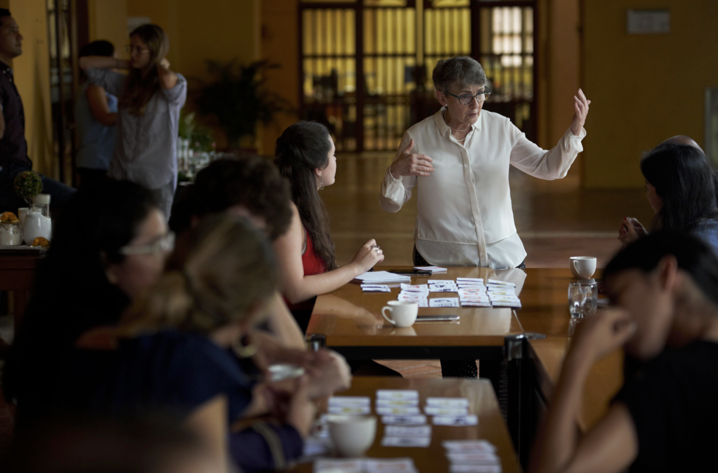 A group of people attending a meeting or workshop in a conference room, with some taking notes, and an older woman standing and speaking to the group.