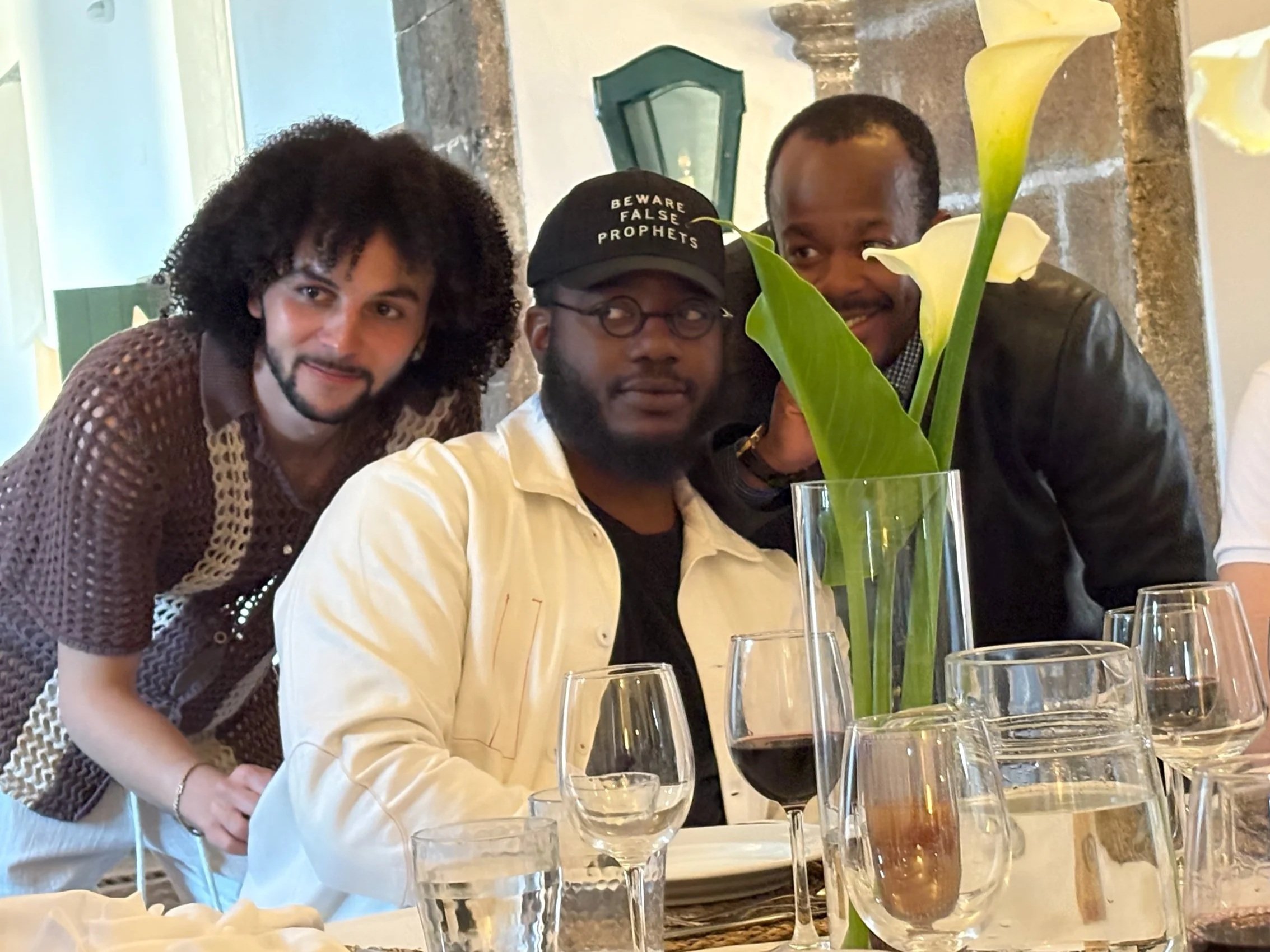 Three men posing together at a social gathering, with a tall white flower in a glass vase in the foreground. The background shows a kitchen or dining area with marble countertops.