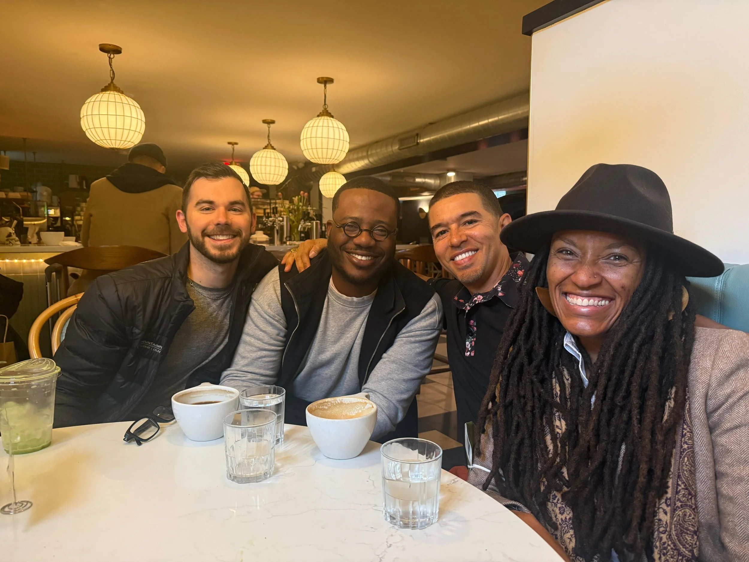 Four smiling friends sitting together at a table in a cozy restaurant, with drinks and cups of coffee or tea in front of them, enjoying a social meal during the evening.