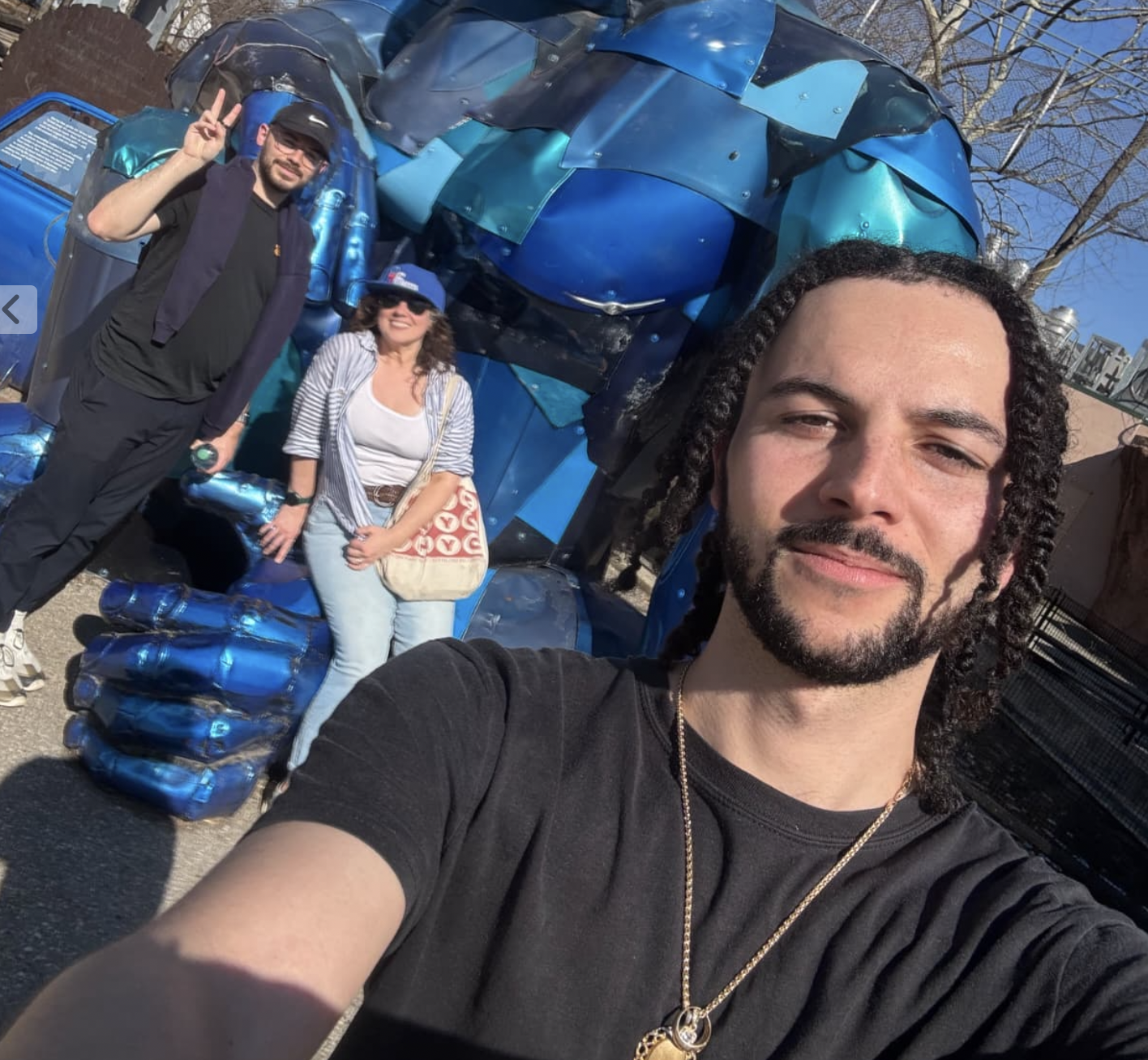 A group of three people taking a selfie in front of a large blue robot sculpture. The person in the foreground has curly hair and is smiling. The other two are standing behind, one making a peace sign. The sculpture resembles the Hulk.