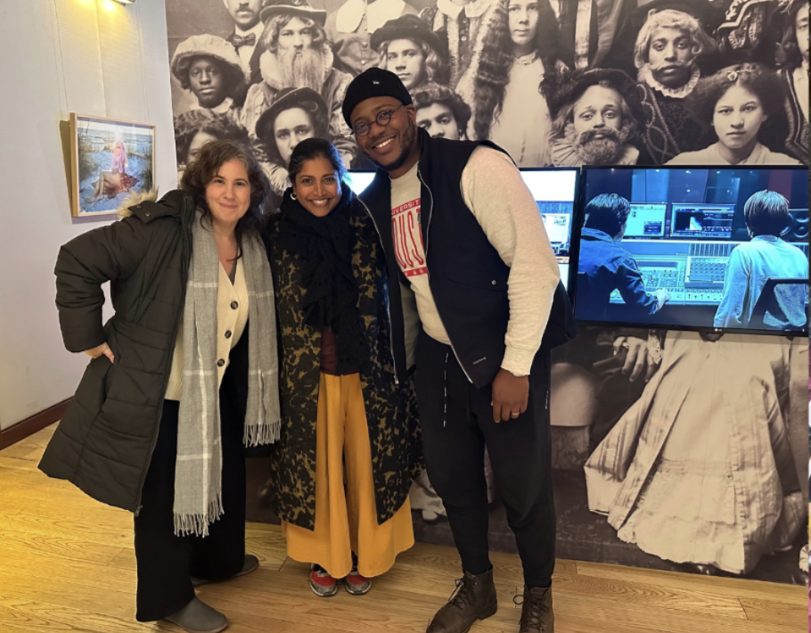 Three people smiling and standing together in an art gallery with a large black-and-white historical photo in the background, and two screens showing editing software.