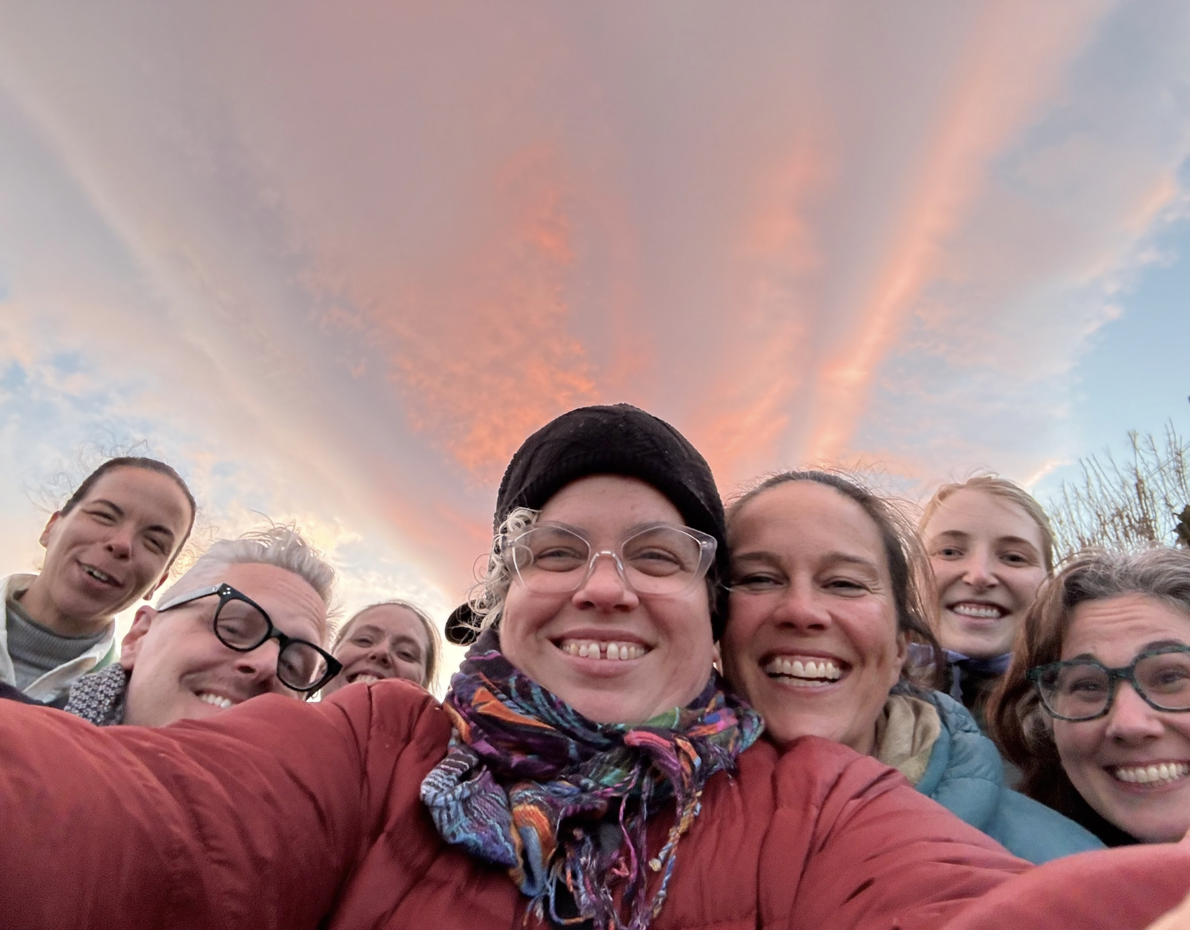 Group of smiling people taking a selfie outdoors during sunset, with a colorful sky and clouds in the background.