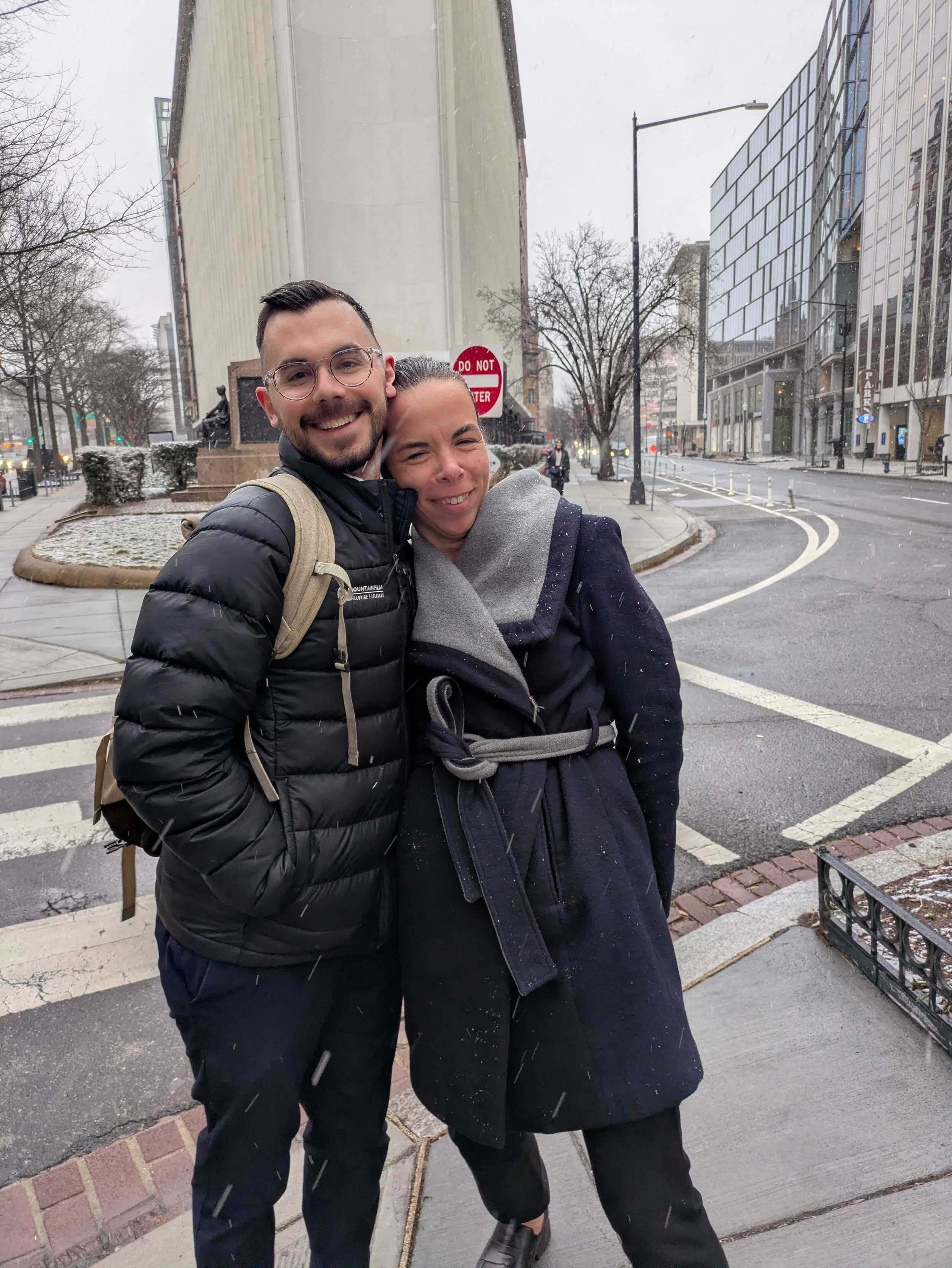 Two smiling people, a man and a woman, hugging outdoors in a city street during winter with snow. The man is wearing glasses and a black jacket with a backpack, and the woman is wearing a dark coat and scarf. There are trees, buildings, and a street 