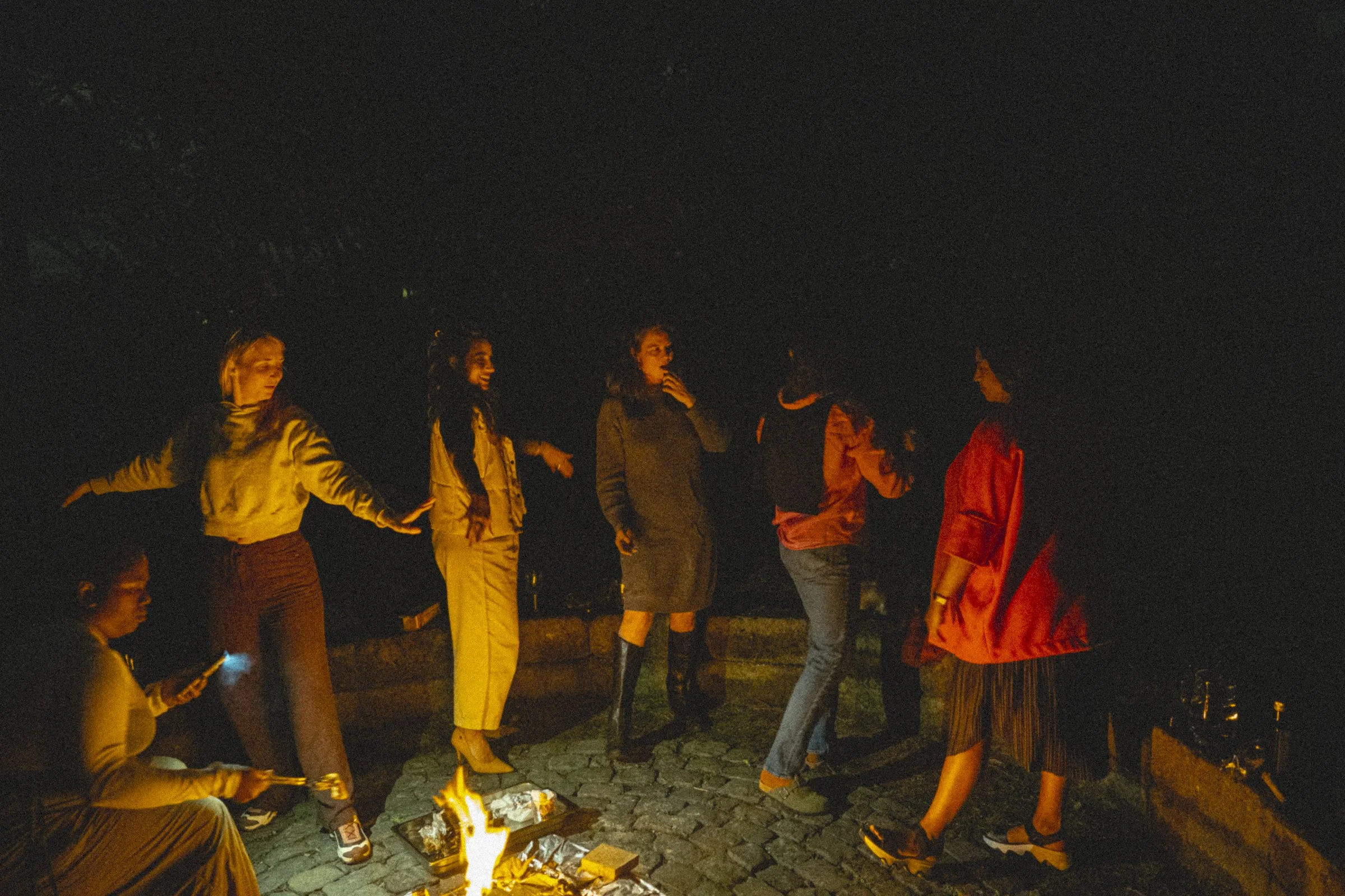 A group of people gathered around a campfire at night, some standing and some sitting, chatting and enjoying the outdoor setting.