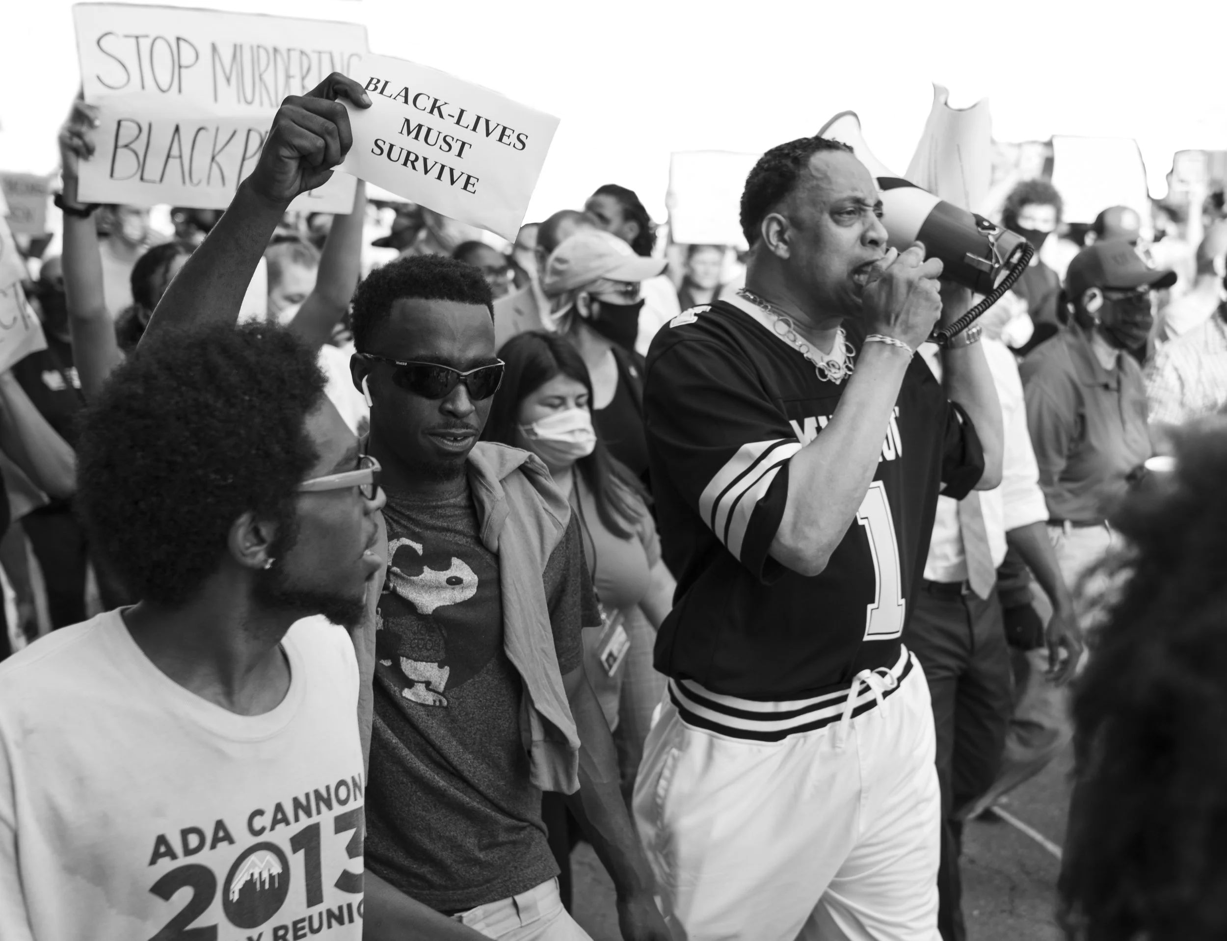 A group of people at a protest or rally, many wearing masks and sunglasses, holding signs about Black Lives Matter and racial justice, with a man speaking into a megaphone.