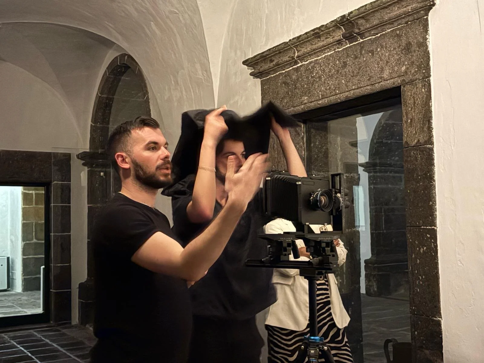 Two people are setting up a camera on a tripod in a room with stone archways and a fireplace, with one person adjusting the camera and the other adjusting their hood.