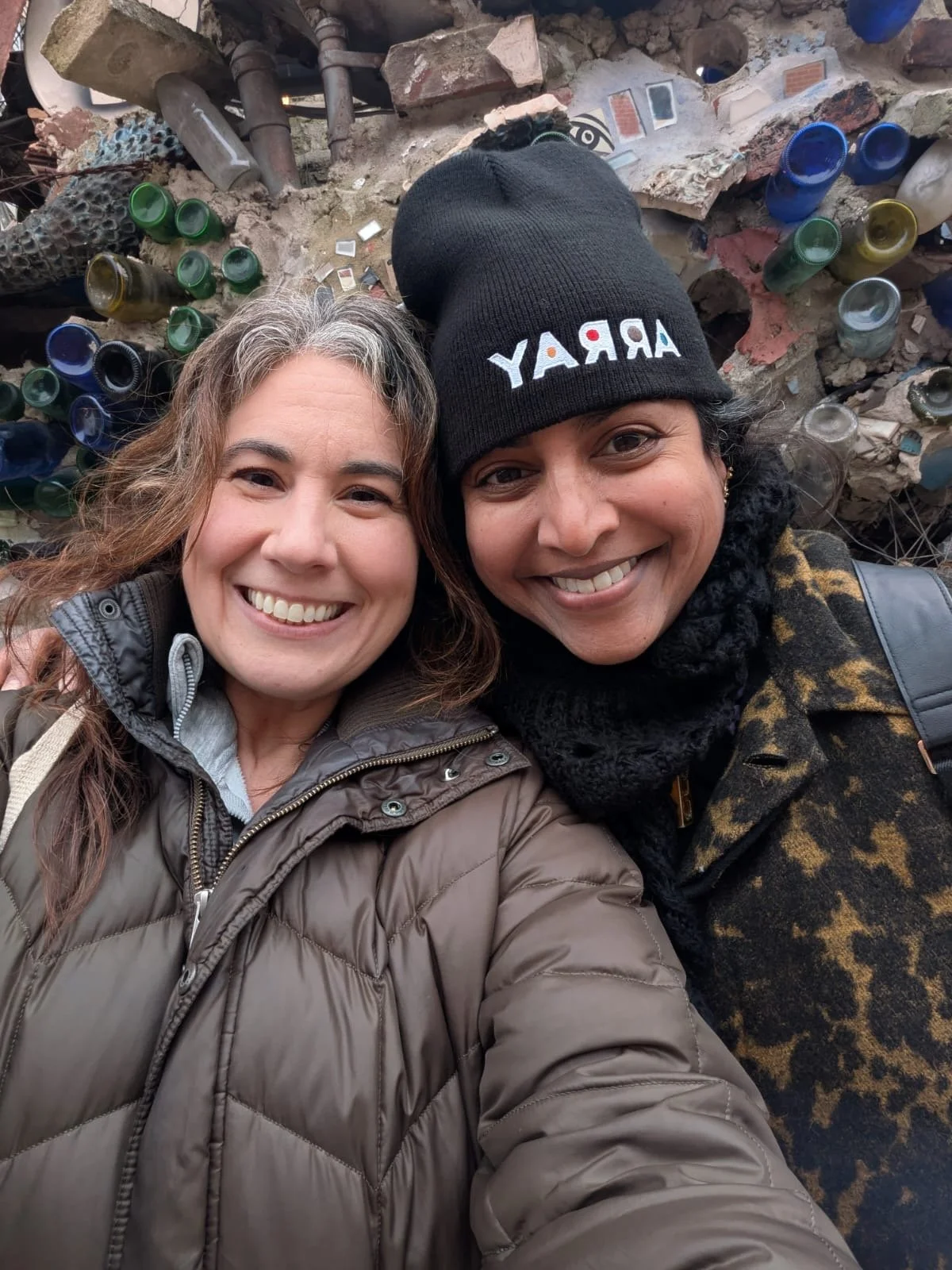 Two smiling women taking a selfie at the Trash Museum, with a wall of recycled glass bottles and debris behind them.