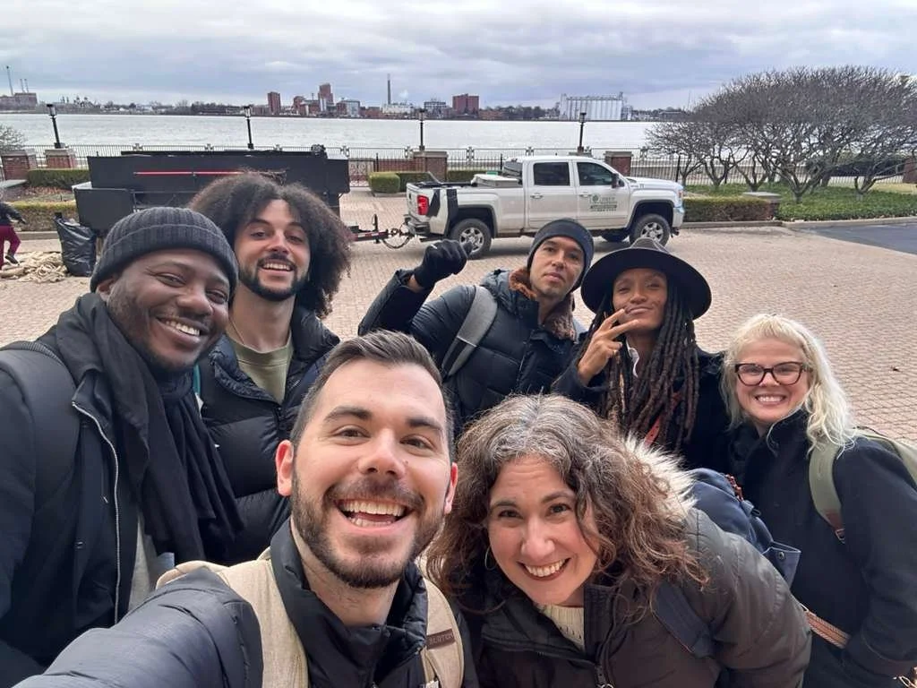 A group of seven diverse people smiling and posing for a selfie outdoors near a waterfront with city buildings in the background.