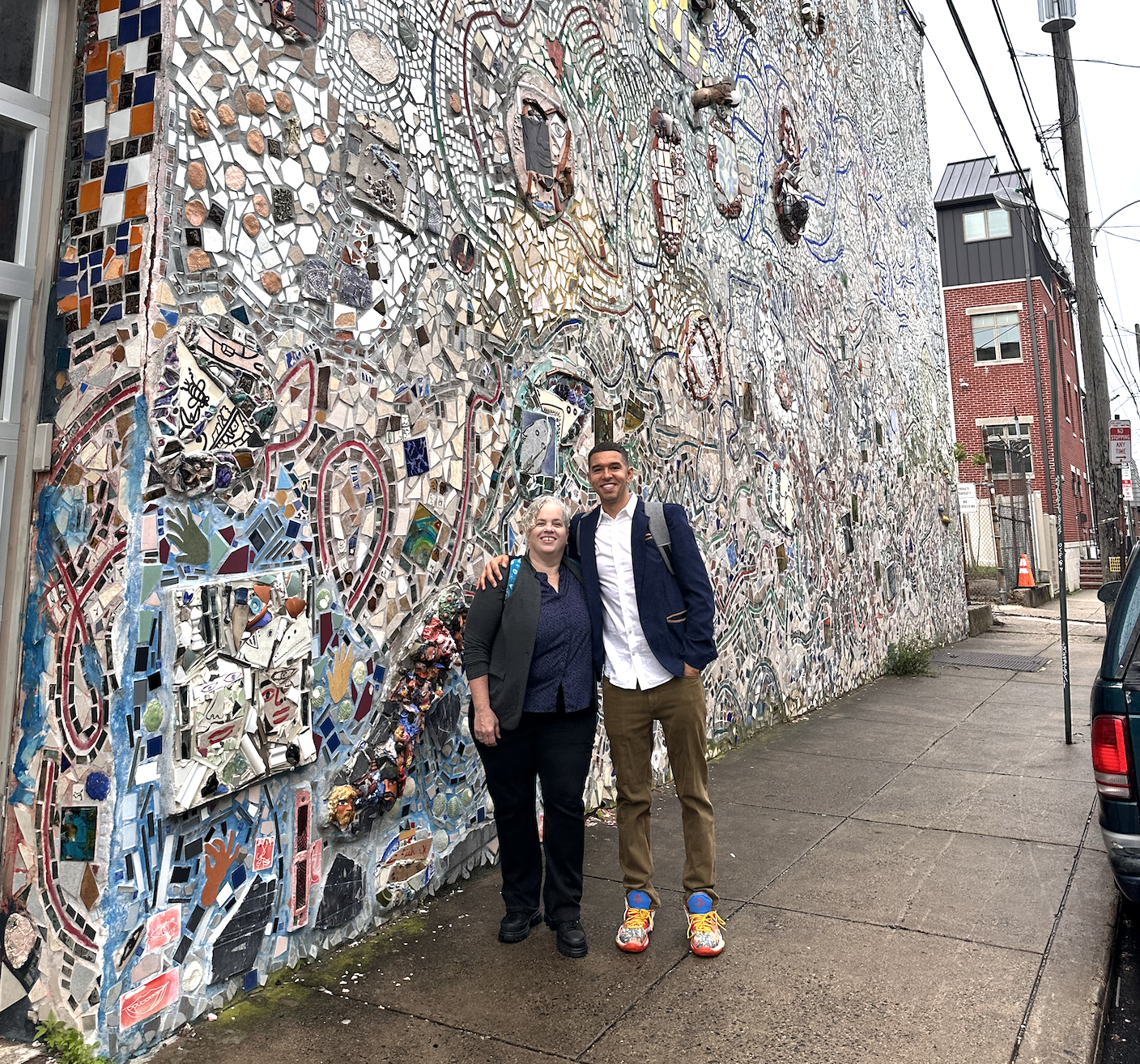 Two people standing in front of a colorful mosaic wall on a sidewalk in an urban area. The wall includes various pieces of broken tiles and artistic decorations. The woman has gray hair, is wearing a black blazer and dark pants, and is smiling with h
