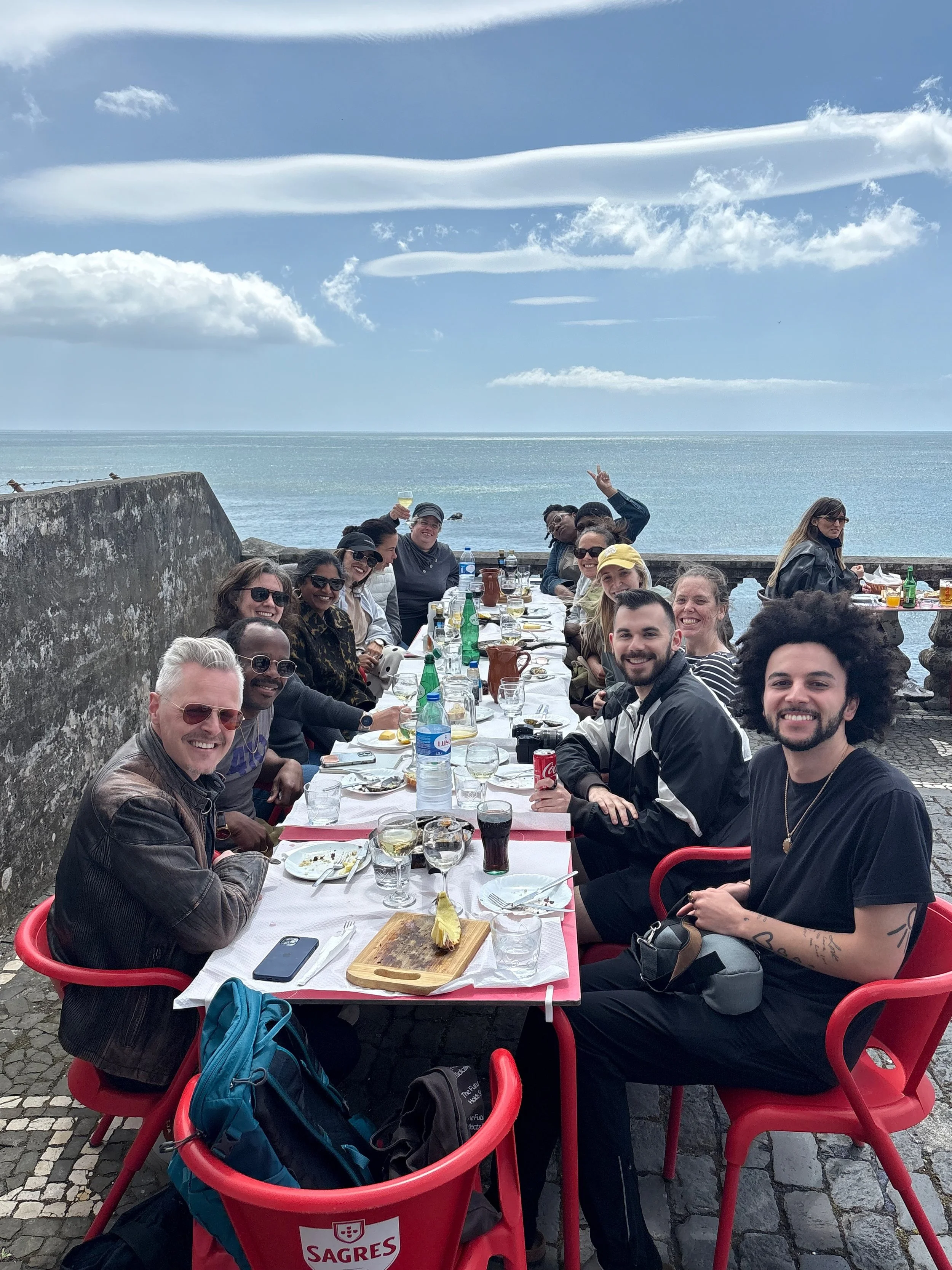 A group of people enjoying a meal outdoors by the water, sitting around a long table with food and drinks, under a partly cloudy sky.