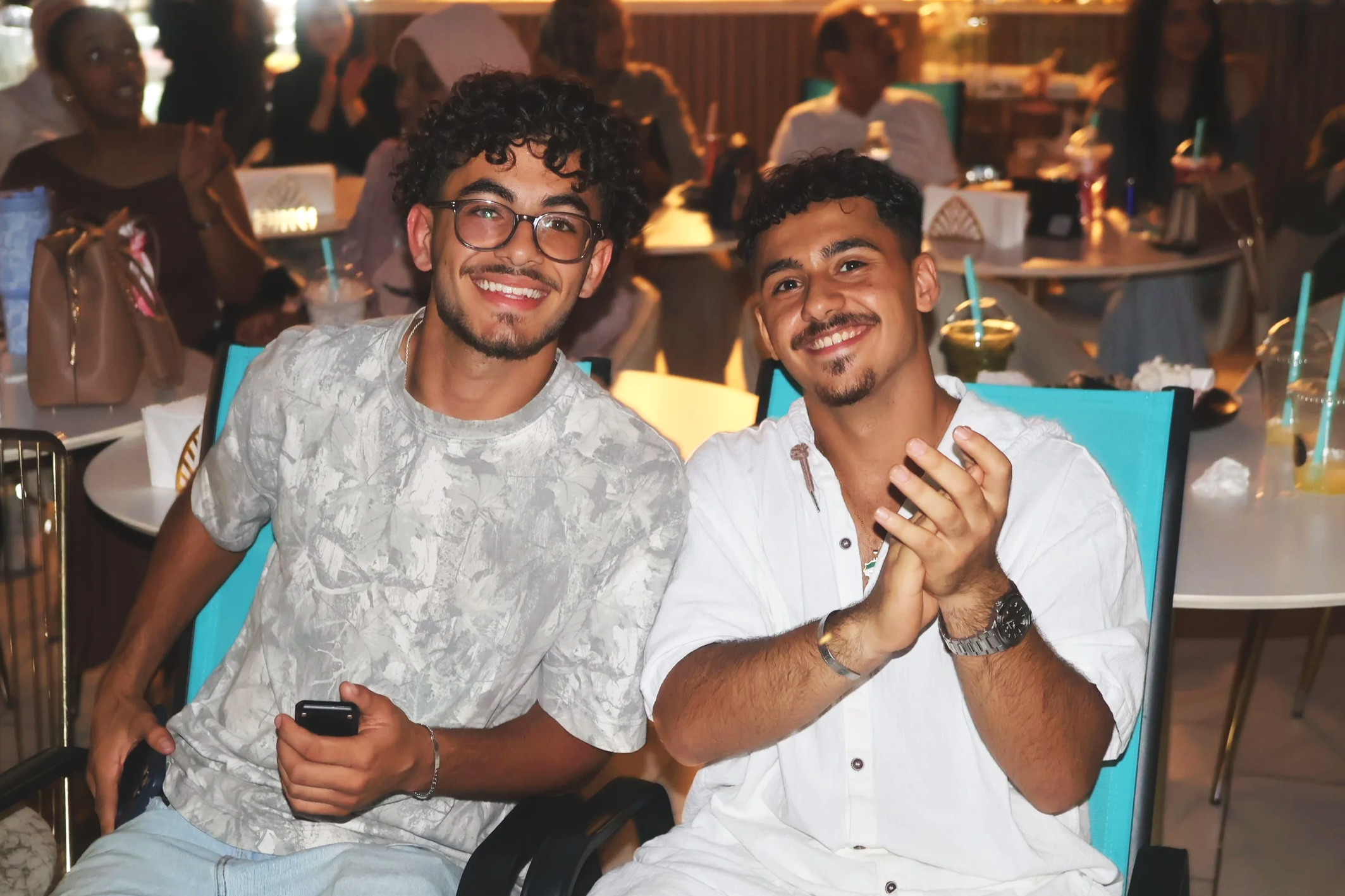 Two young men with curly hair and smiling faces sitting close together at a social event, with other people in the background at a restaurant or cafe.