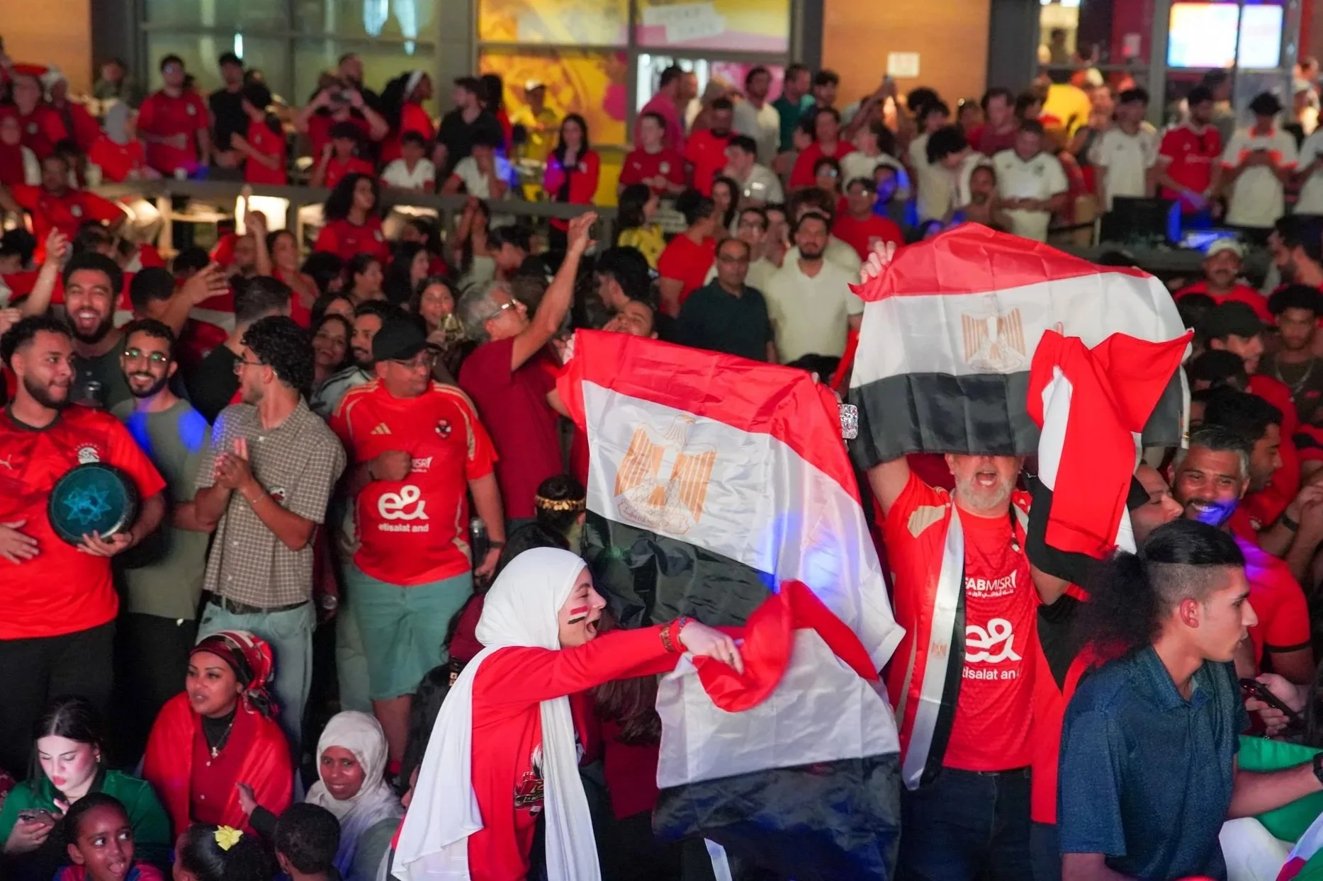 A large crowd of people gathered indoors celebrating Egyptian national pride. Many are wearing red, black, and white clothing, and some are holding Egyptian flags. The atmosphere is festive and joyful, with some people waving flags and others smiling