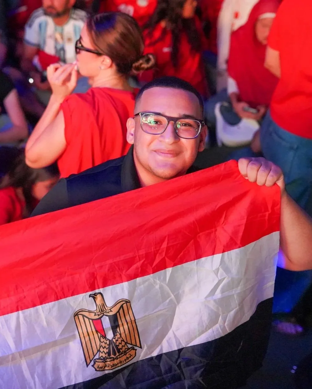A young man wearing glasses, smiling, holding an Egyptian flag at a crowded event.