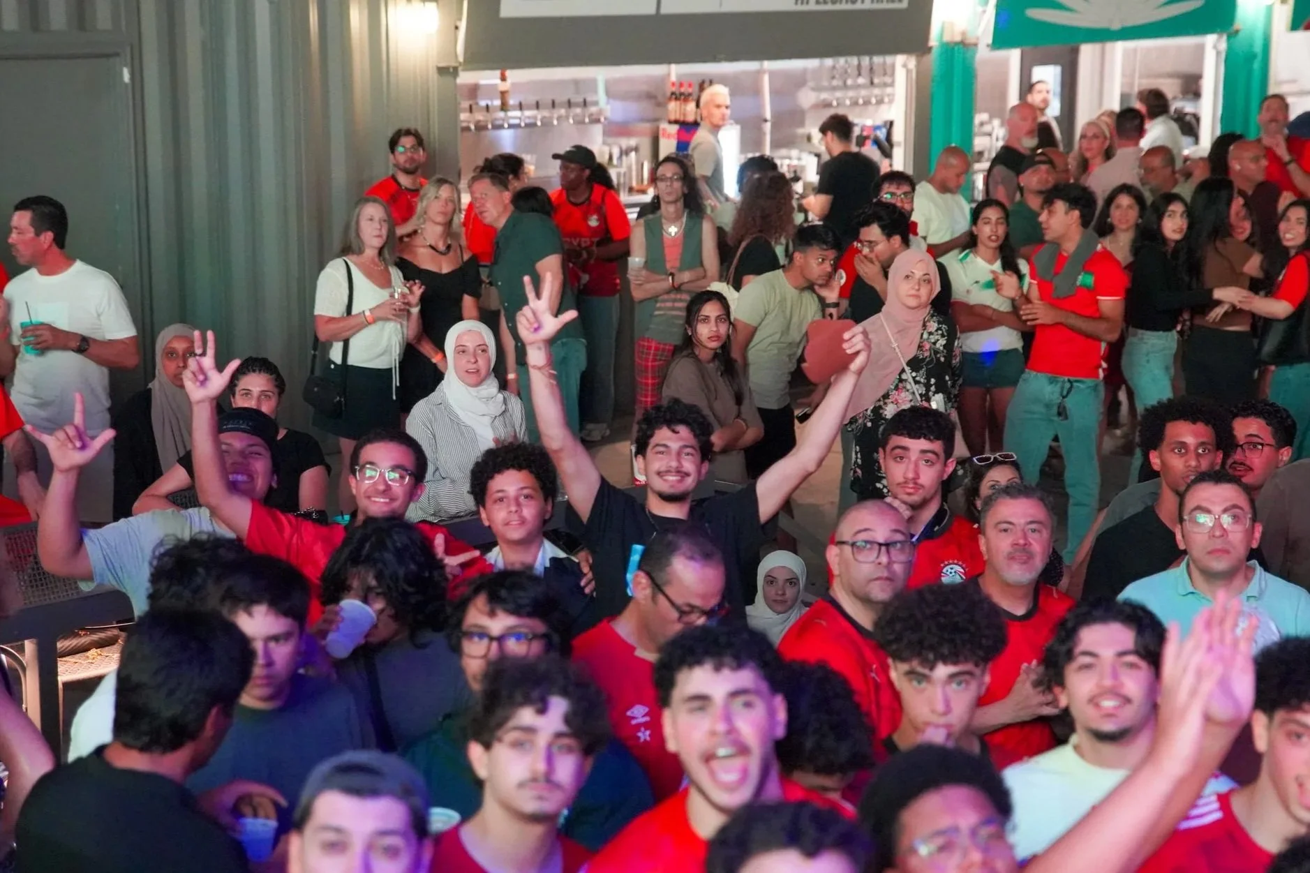 A large group of diverse young people gathered indoors, some wearing red clothing, celebrating and socializing at a crowded event or party.