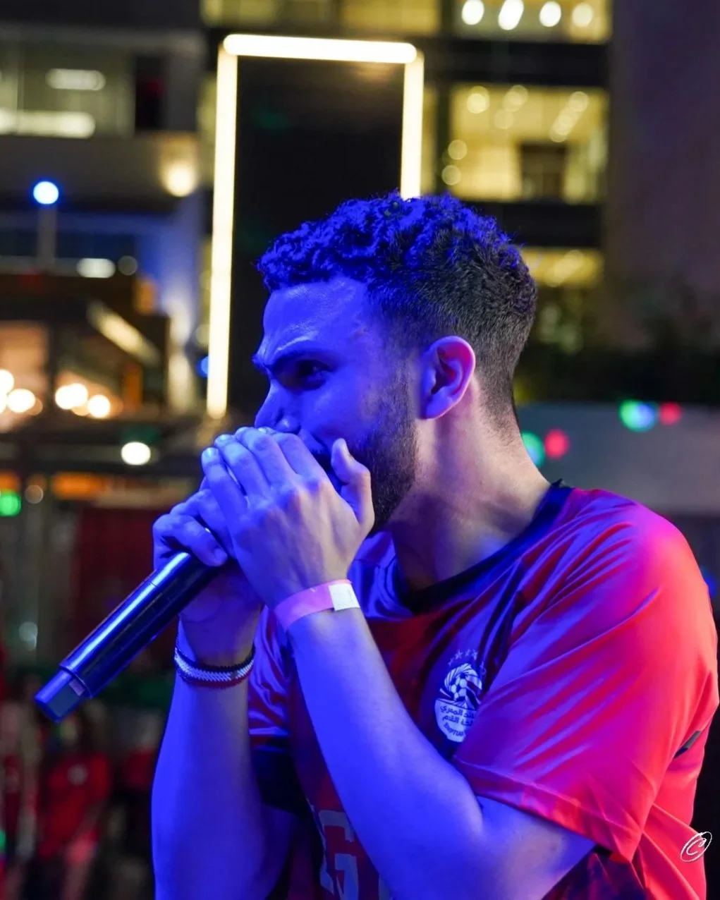 A man with a beard and short curly hair singing into a microphone in a dimly lit venue with colorful lights and modern decor.