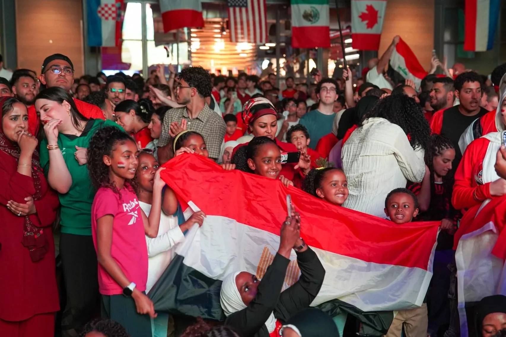 Crowd of diverse people celebrating, some holding a large flag, with international flags hanging overhead in the background.