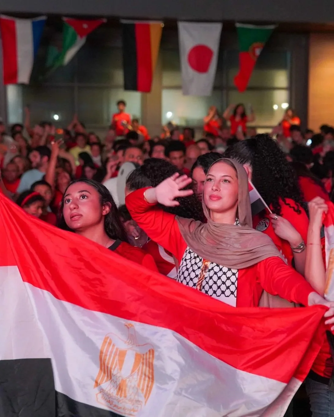 A diverse crowd of women and men celebrating with Egyptian flags at an indoor event, with colorful banners hanging from the ceiling.