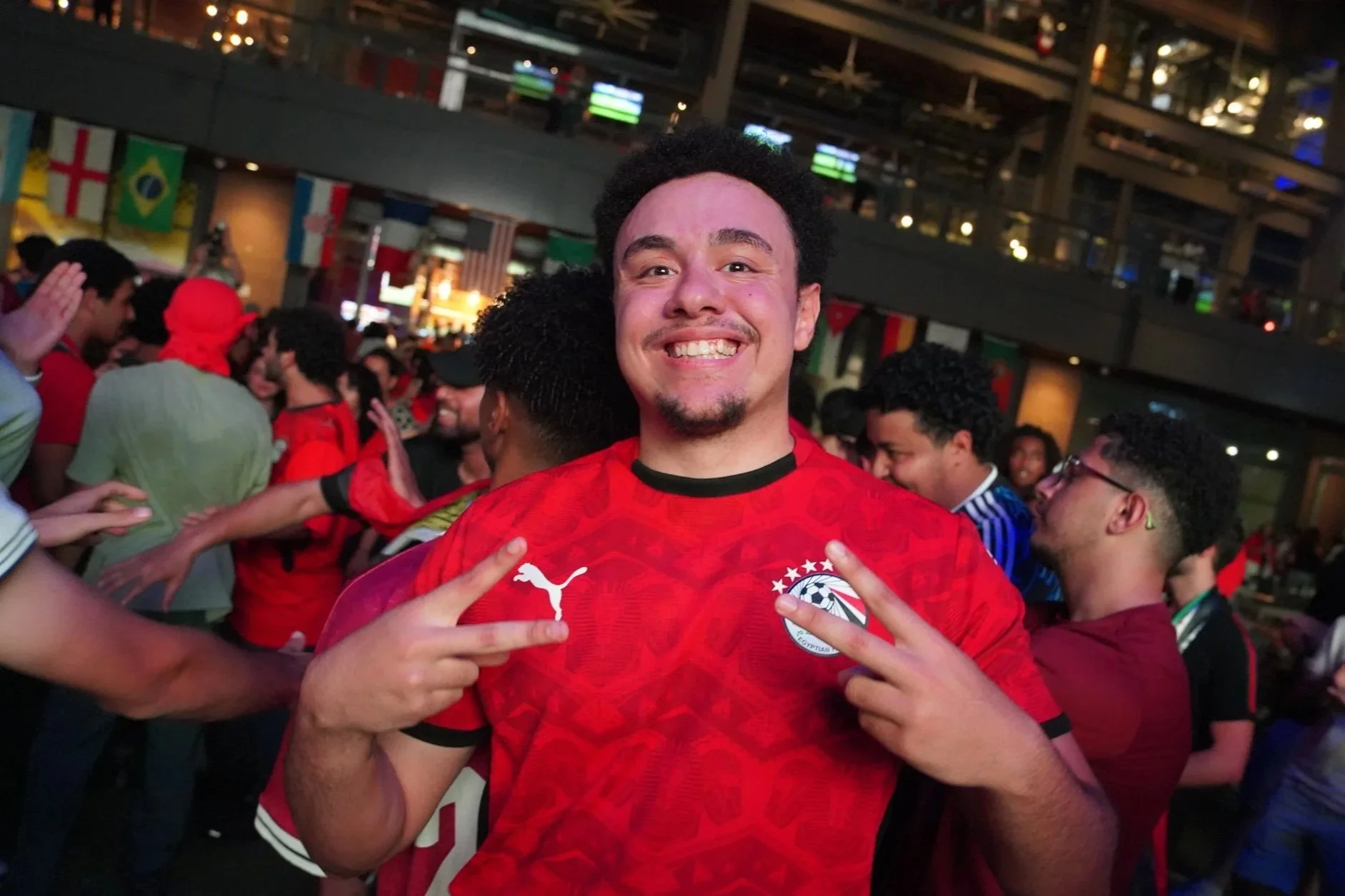 Smiling man wearing a red sports jersey with a Puma logo, pointing at the camera, surrounded by people at a lively indoor event with flags and banners in the background.