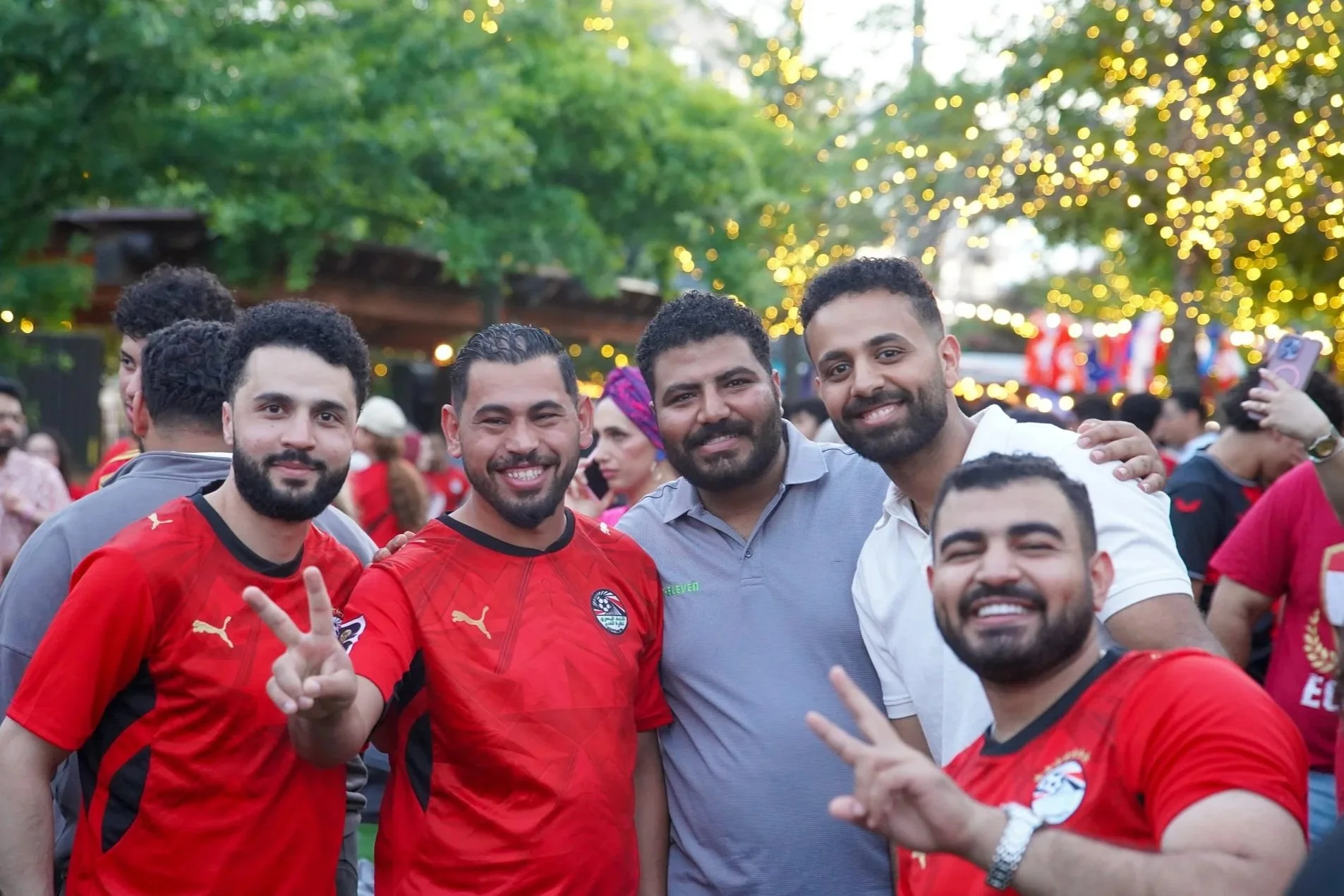 Group of smiling men, some wearing red sports jerseys, gathering outdoors with trees and string lights in the background.