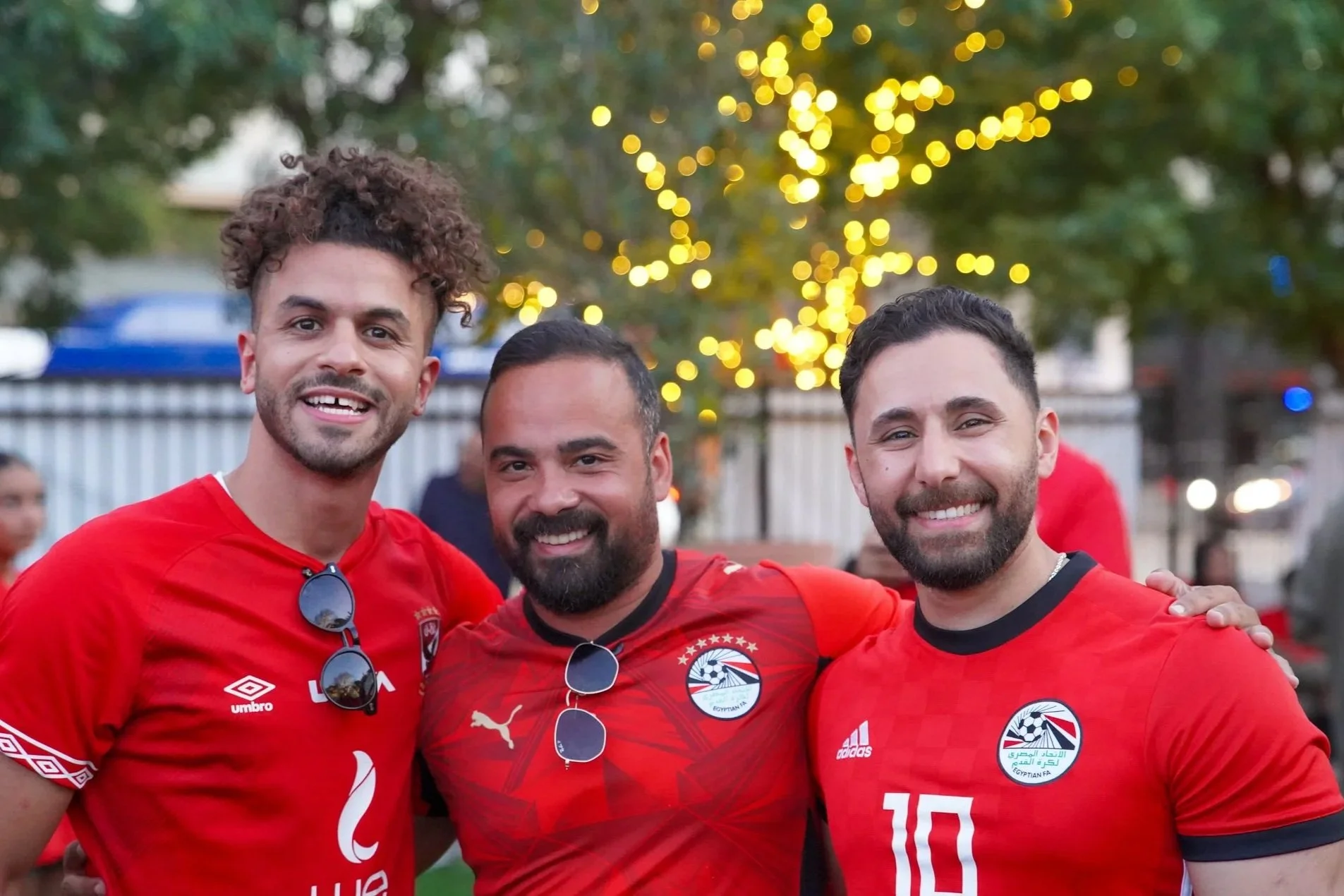 Three men wearing red soccer jerseys with Egyptian Football Association badges, smiling and standing close together at an outdoor event during the evening, with blurred background and decorative lights.