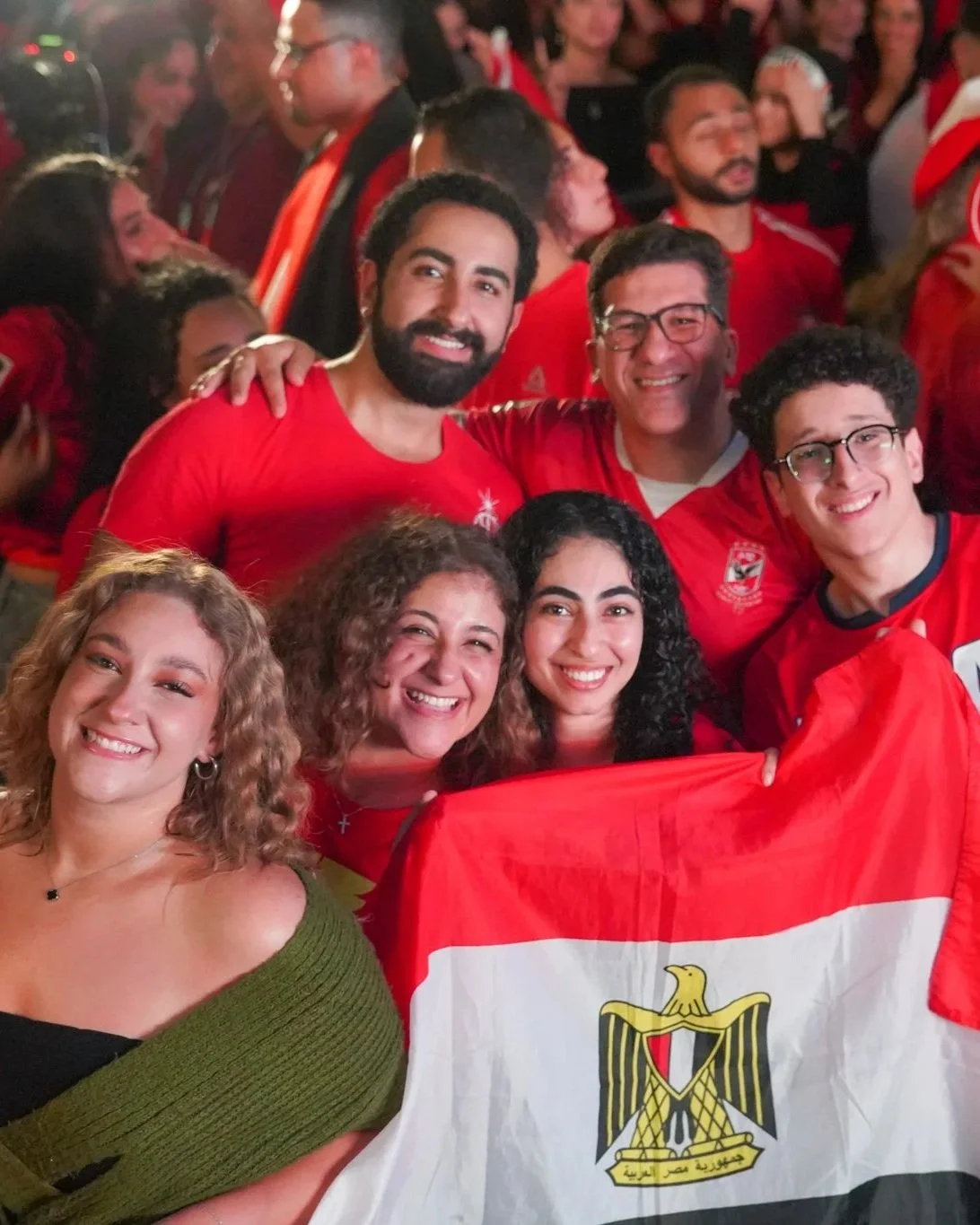 Group of diverse people celebrating, holding an Egyptian flag, many dressed in red, smiling, in a lively indoor setting.