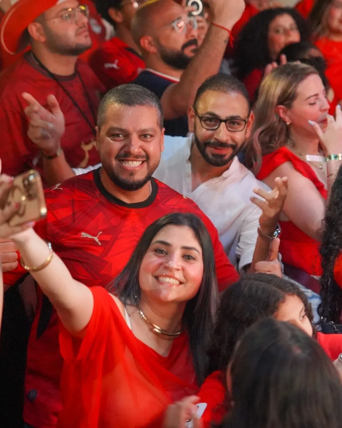 A group of people at a celebration, wearing red, smiling, and some making gestures, with the focus on a young woman in the foreground taking a selfie.