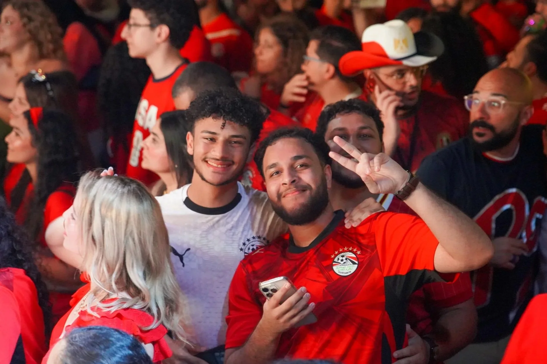 A group of people celebrating, many wearing Red Egypt football jerseys, some smiling at the camera, others talking or looking around, in a crowded festive atmosphere.