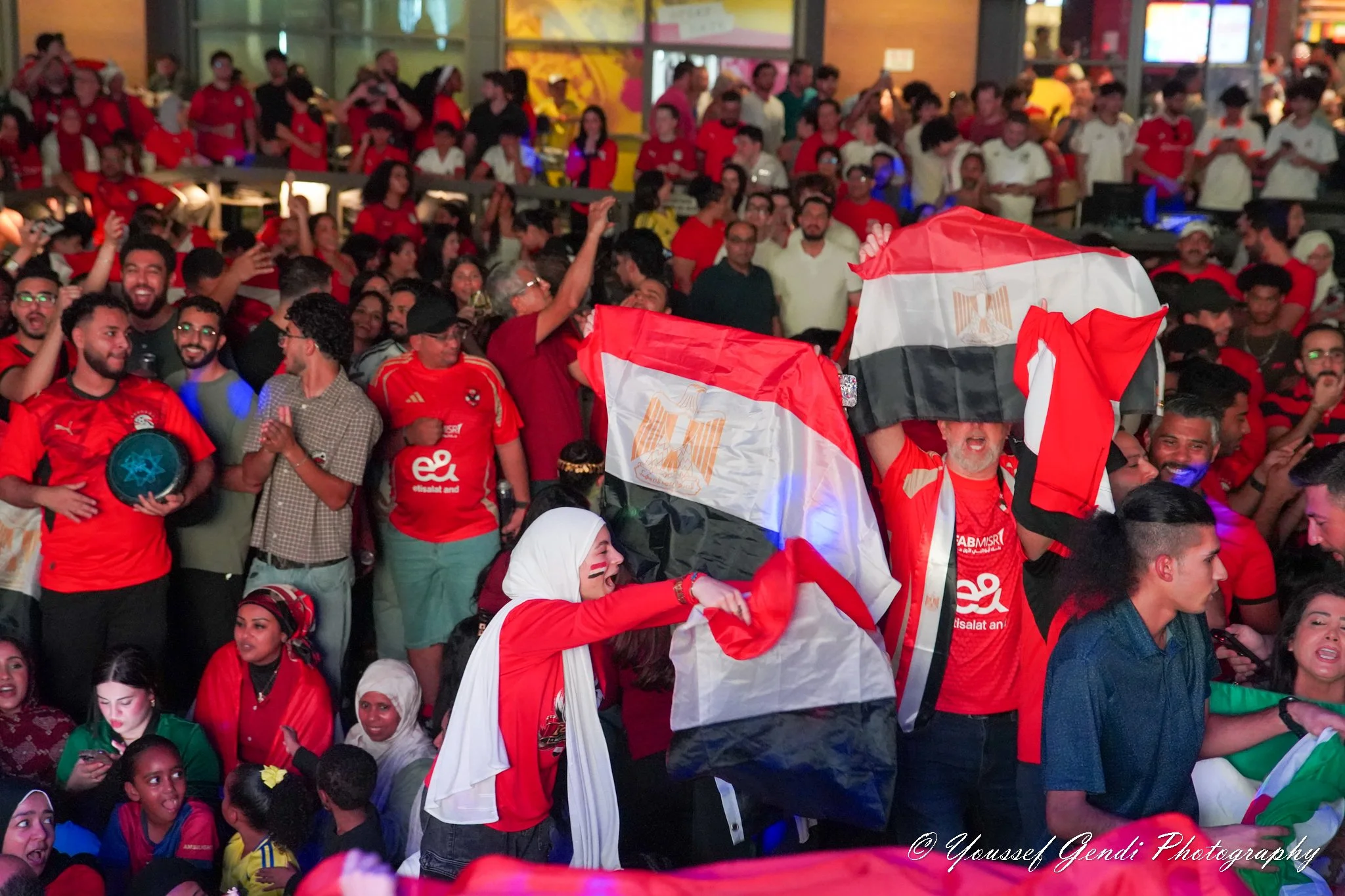 A large crowd of Egypt Fans celebrating, dressed in red Egyptjerseys, some waving Egyptian flags, inside Legacy Hall Plano with glass and brick walls.