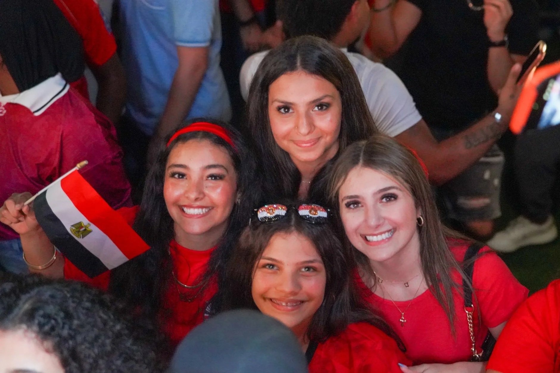 A group of women smiling and celebrating, with one woman holding a small Egyptian flag. They are gathered closely together, and some are wearing red clothing and accessories, suggesting they are celebrating an event or national pride.