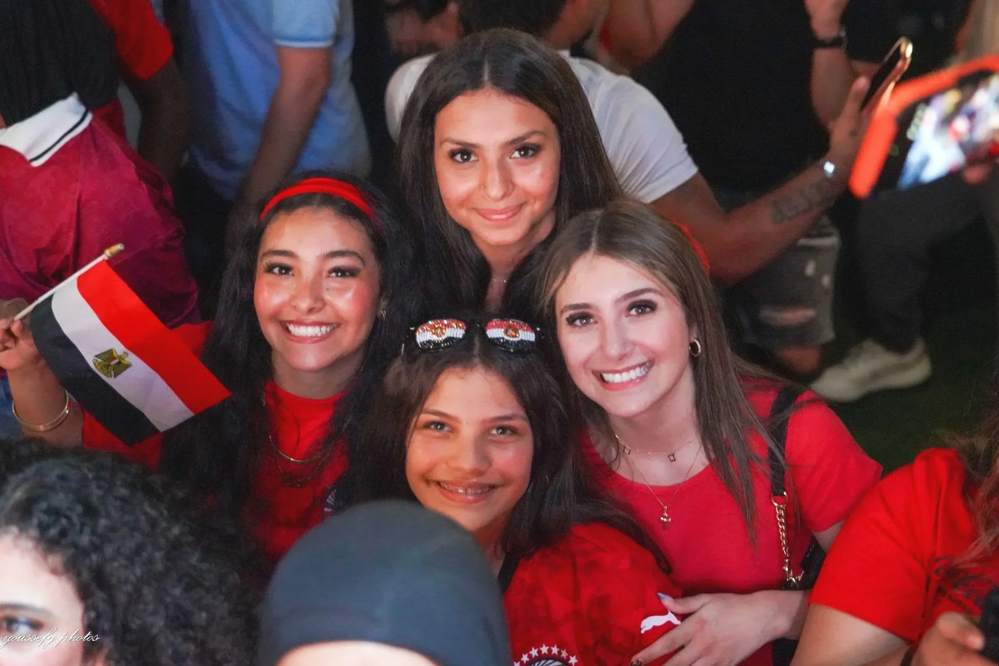 Group of young women celebrating, some holding Egyptian flags, in a crowded gathering, wearing red shirts and accessories.