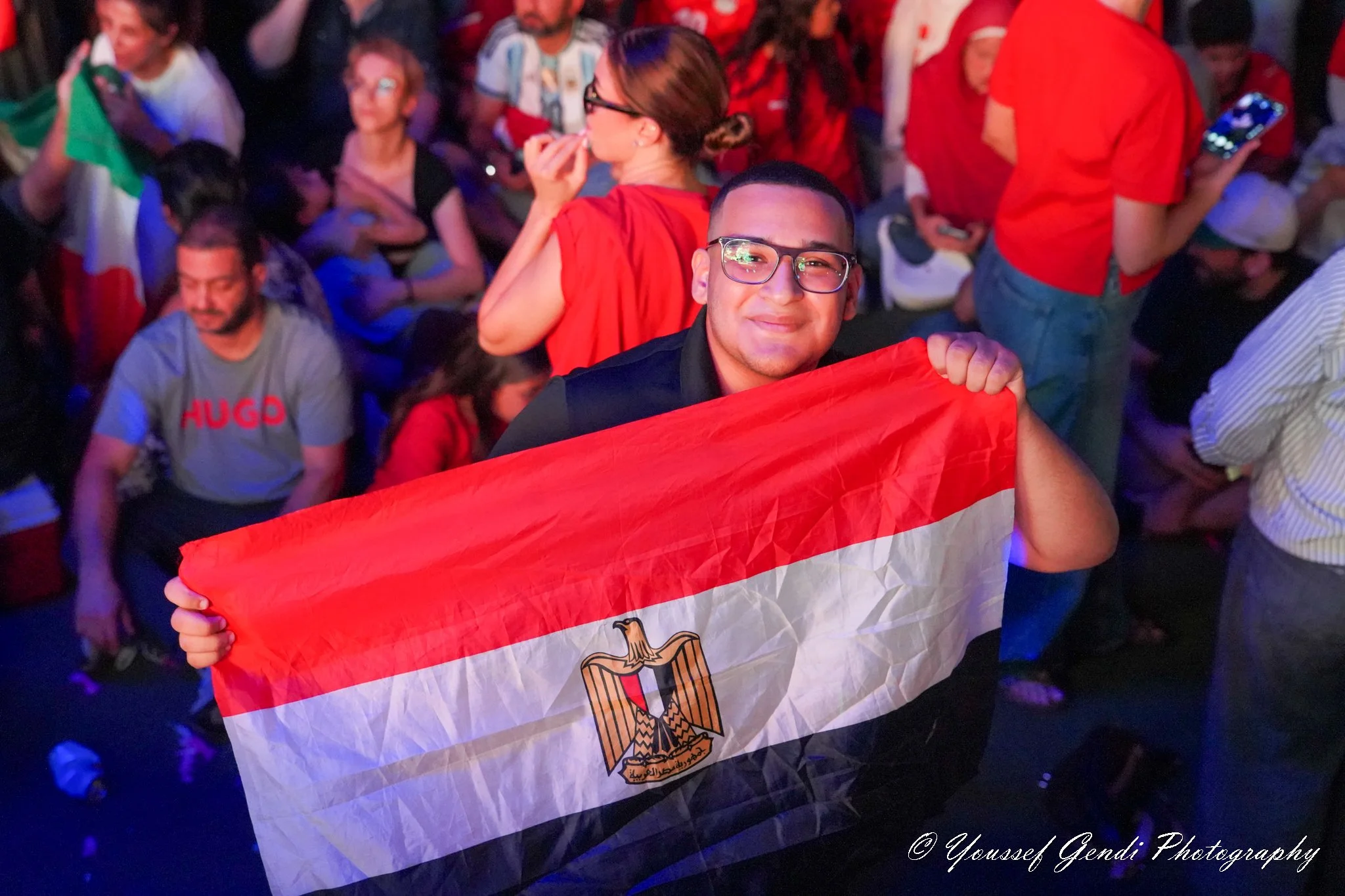 A man smiling and holding an Egyptian flag, with a crowd of people dressed in red around him, some holding phones and flags, at what appears to be a celebration at Legacy Hall in Plano DFW.
