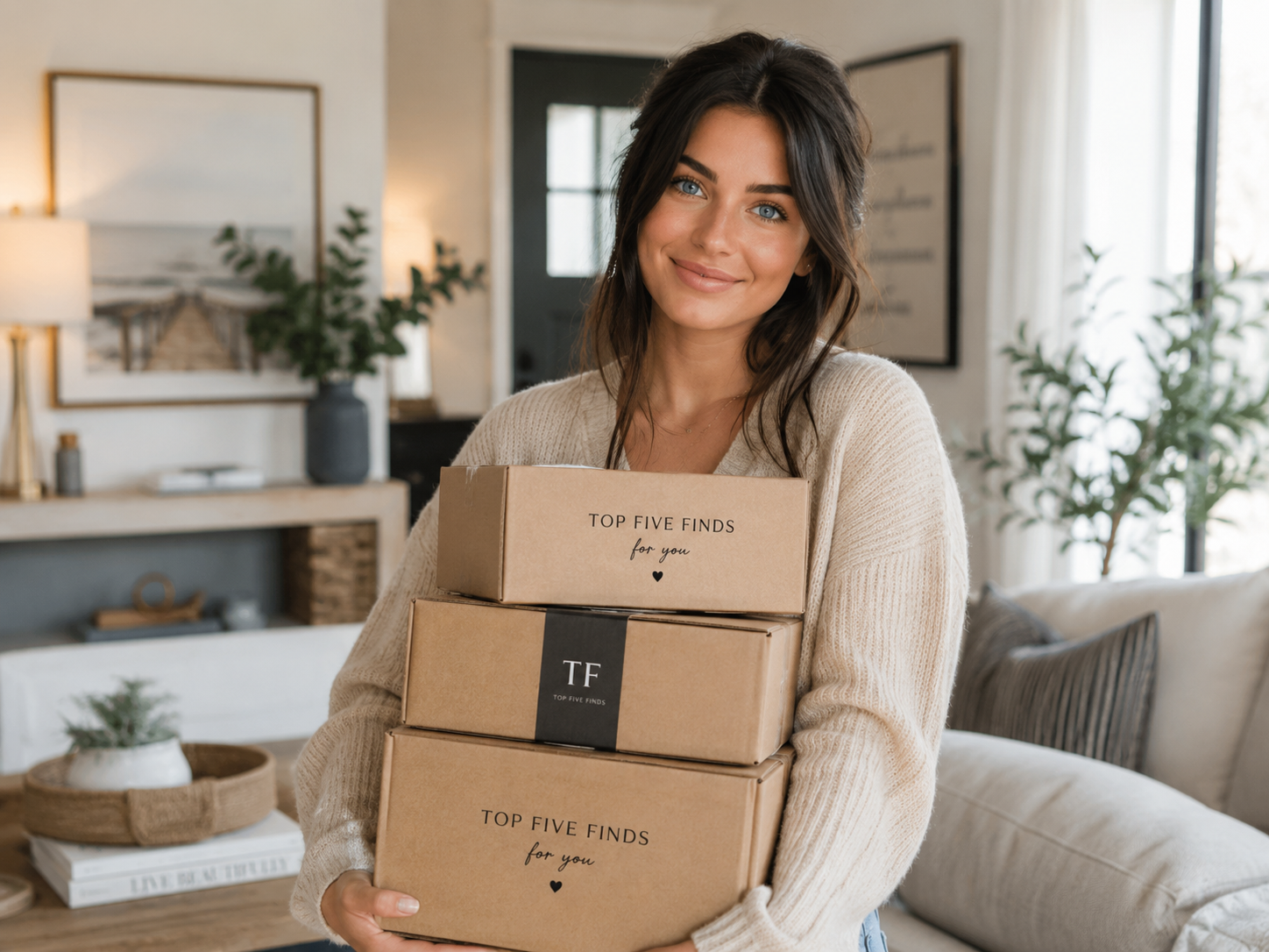 A woman with dark hair and blue eyes smiling while holding three cardboard boxes labeled "Top Five Finds for You" in a cozy living room.