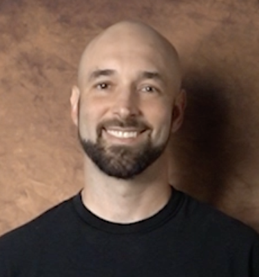 Portrait of a smiling bald man with a beard wearing a black shirt, standing against a brown background.
