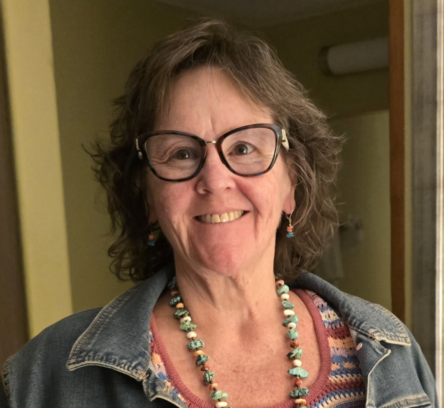 A smiling woman with curly brown hair, wearing large glasses, colorful beaded jewelry, a denim jacket, and a patterned shirt, standing indoors.