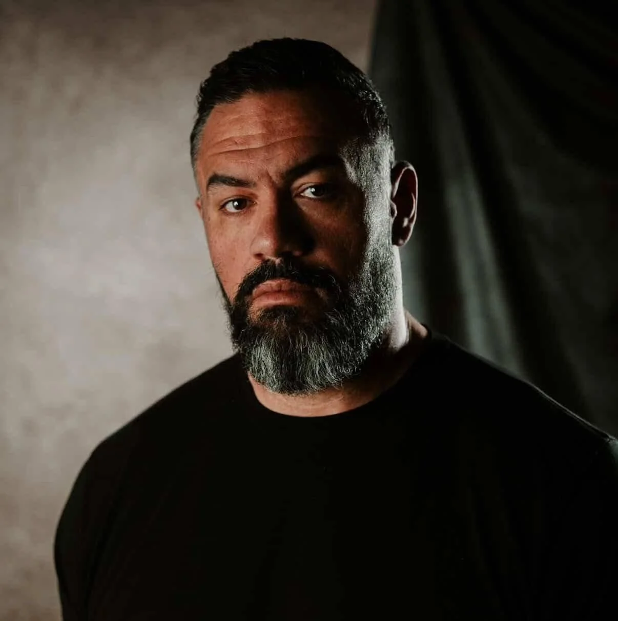 A man with dark hair and a beard, wearing a black shirt, looking directly at the camera with a serious expression.