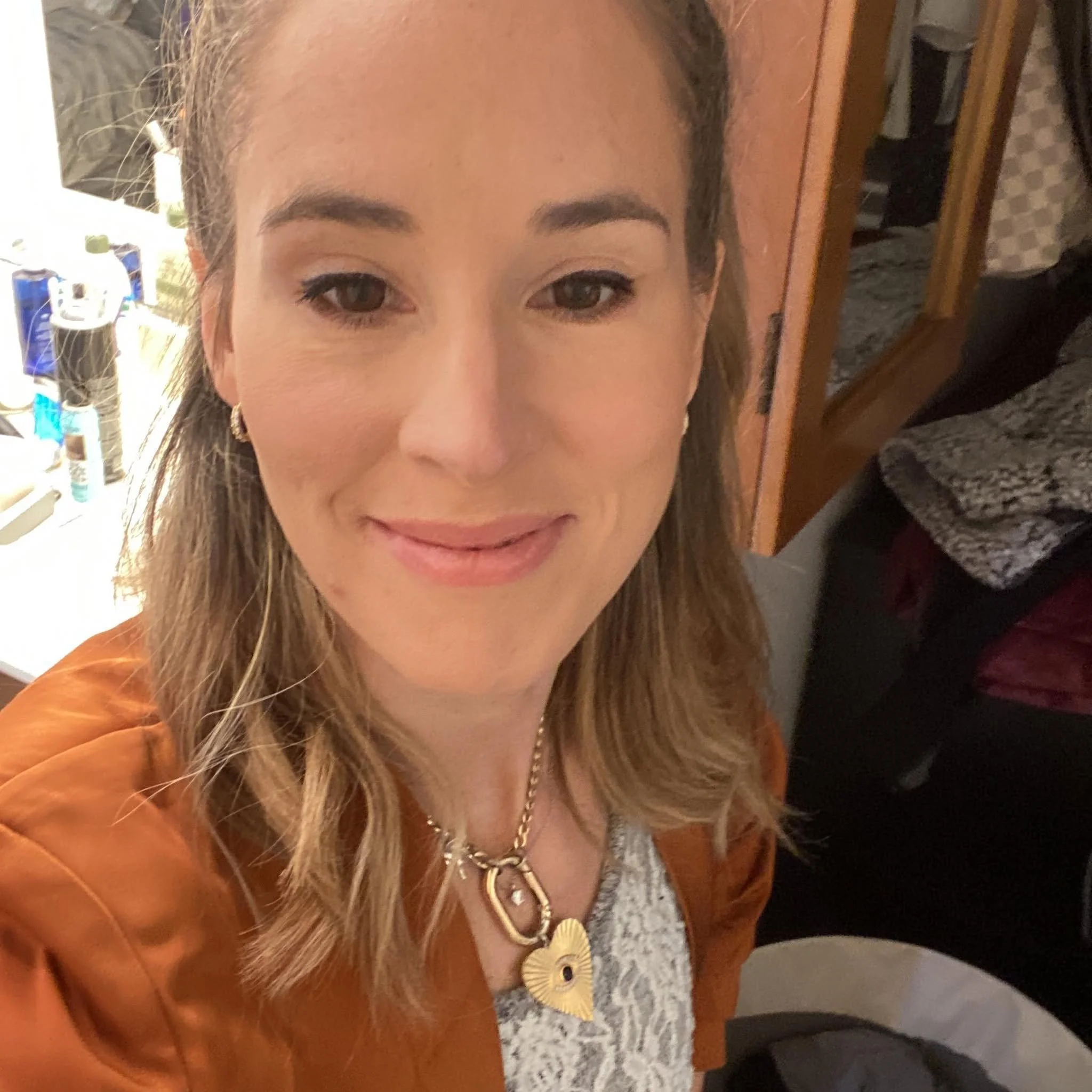 Close-up of a woman's face, showing her eyes, nose, and partial smile, with a necklace featuring a gold fan-shaped pendant, brown hair, and a rust-colored jacket, in an indoor setting.