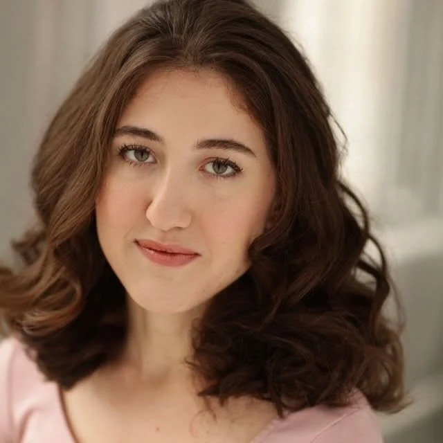 Close-up portrait of a young woman with wavy brown hair and light makeup, looking at the camera with a slight smile, in a softly lit indoor setting.