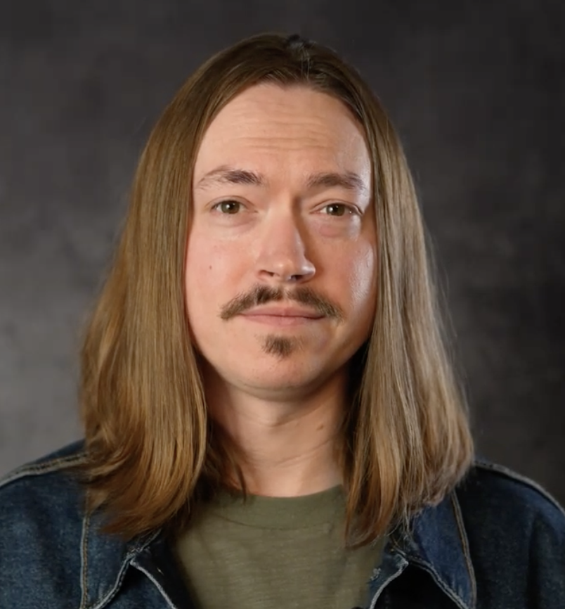A man with long, straight hair, a mustache, and a small goatee, wearing a denim jacket and a green shirt, posing against a dark background.