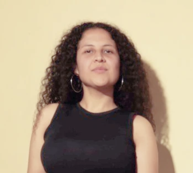 Young woman with curly hair wearing a black sleeveless top and hoop earrings standing against a yellow wall.