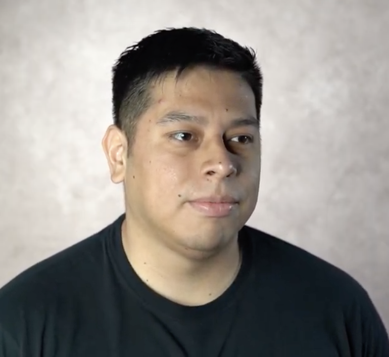 Portrait of a young man with short black hair wearing a black T-shirt, looking slightly to the side against a neutral background.