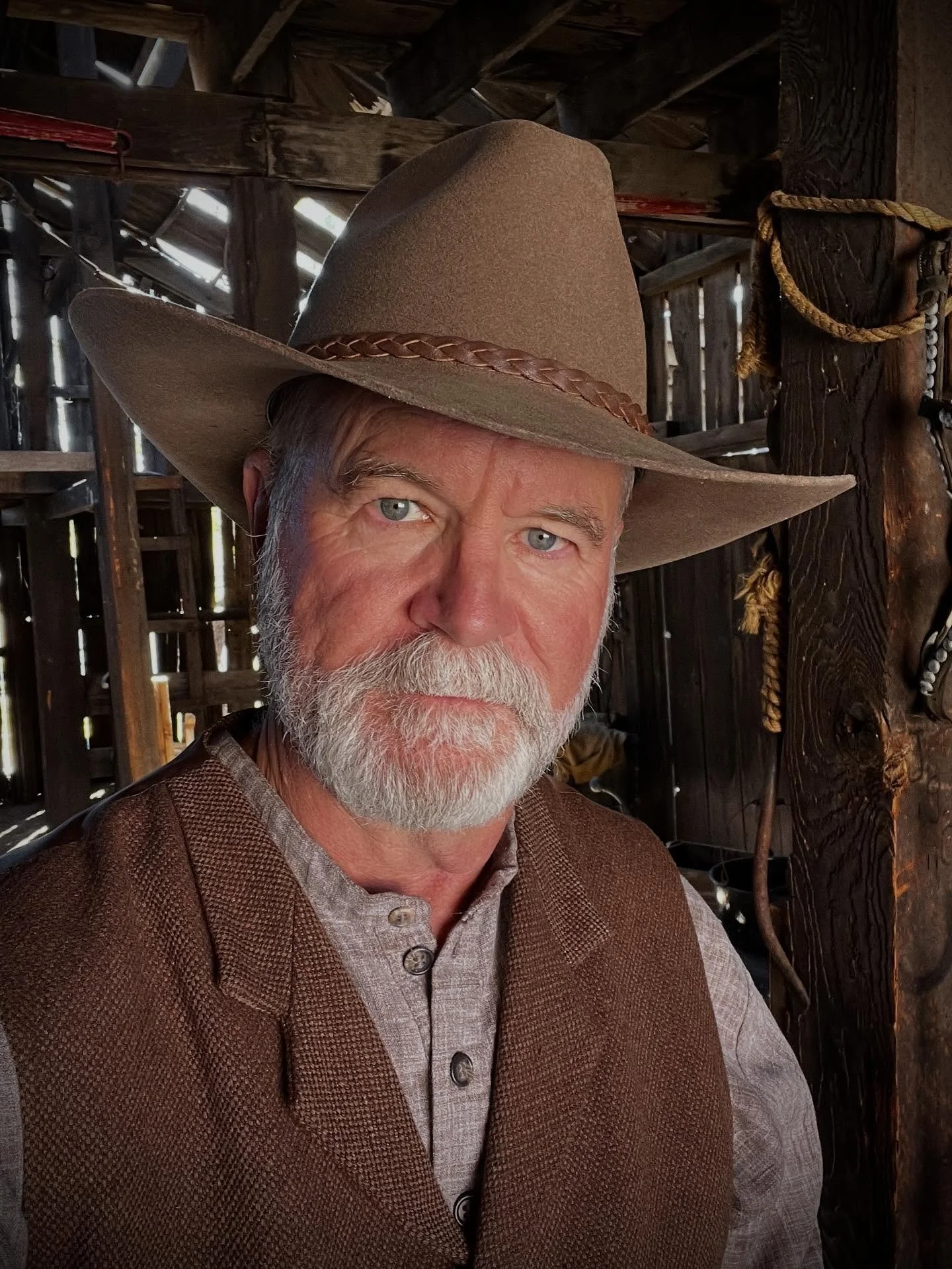 A close-up photo of an older man with a beard and blue eyes, wearing a brown wide-brimmed cowboy hat and a brown vest, sitting in a rustic wooden barn.