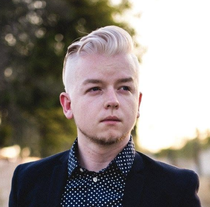 Young man with blonde hair styled back, wearing a dark blazer over a black shirt with white polka dots, standing outdoors with blurred trees in the background.