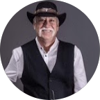 Portrait of a man wearing a cowboy hat, black vest over a white shirt, smiling against a dark background.