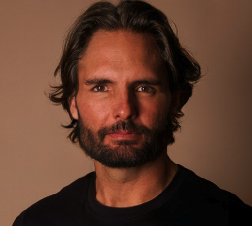 A man with shoulder-length dark hair and a beard, wearing a black shirt, looking directly at the camera with a neutral expression.