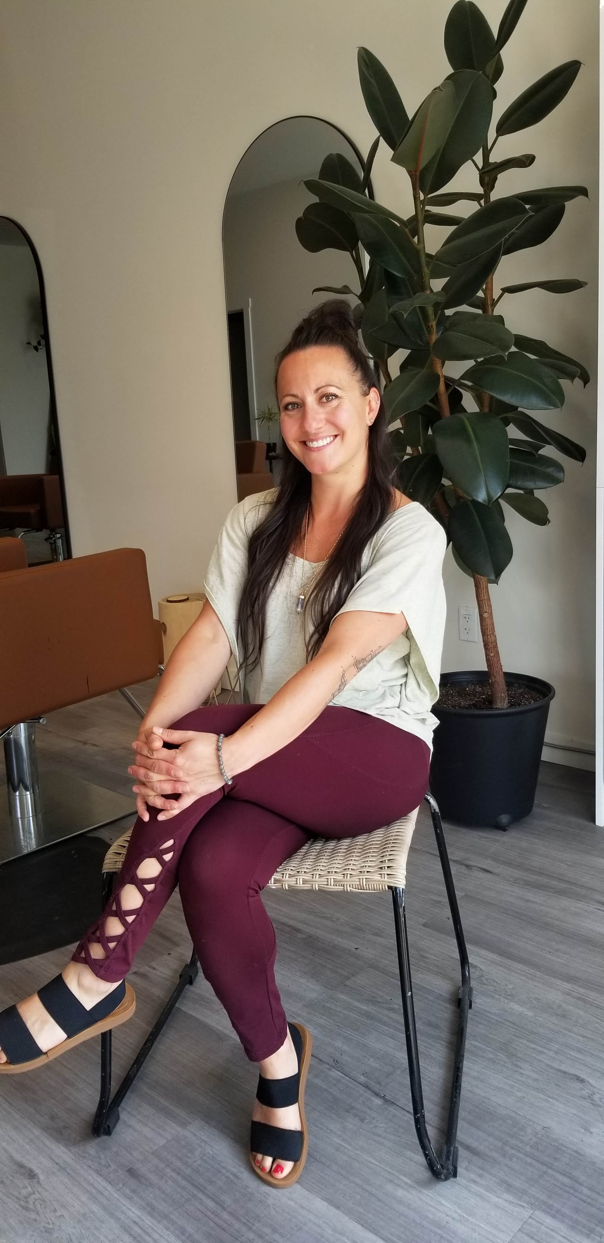 Woman sitting on a chair, smiling, with a large potted plant behind her in a modern interior.
