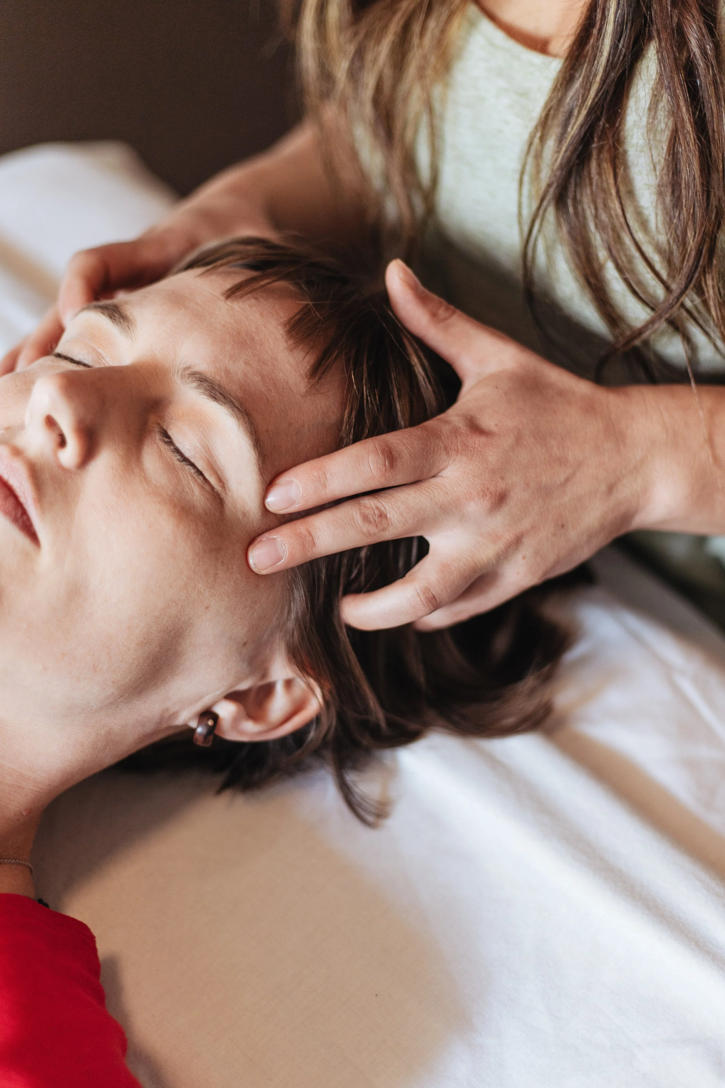 Close-up of a woman giving a neck massage to a woman with short brown hair. The woman receiving the massage has her eyes closed and is lying down on a surface. The emphasis is on the hands and neck area.